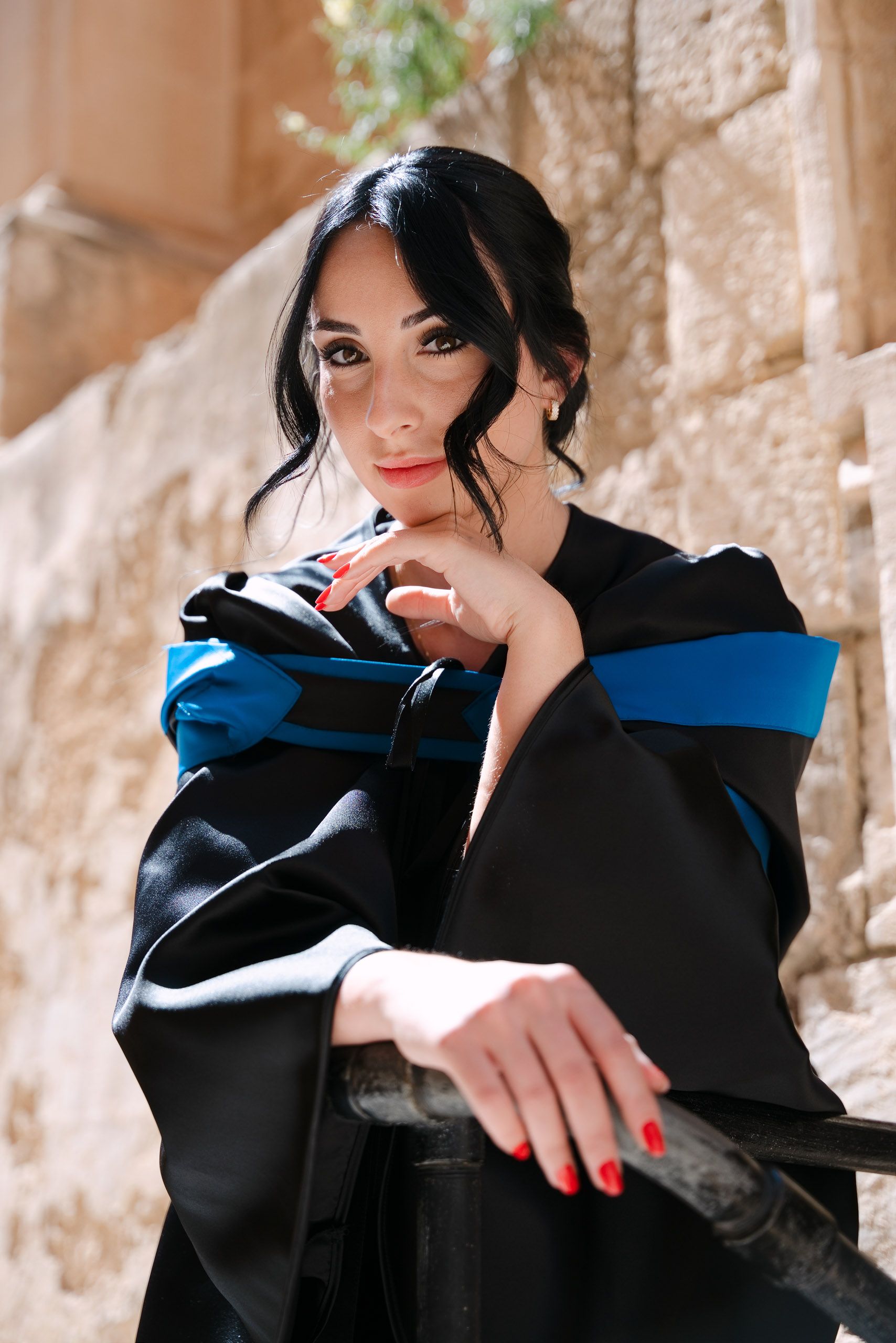 Close-up portrait of a female graduate in a blue-trimmed gown leaning against a railing in the limestone streets of Mdina, Malta.