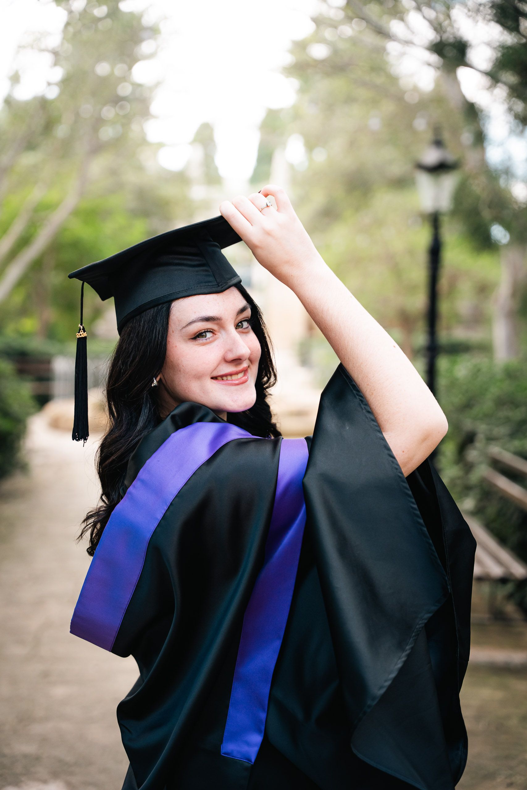 Graduate in a black gown and purple hood smiling while adjusting her cap at Romeo Romano Gardens in Malta.