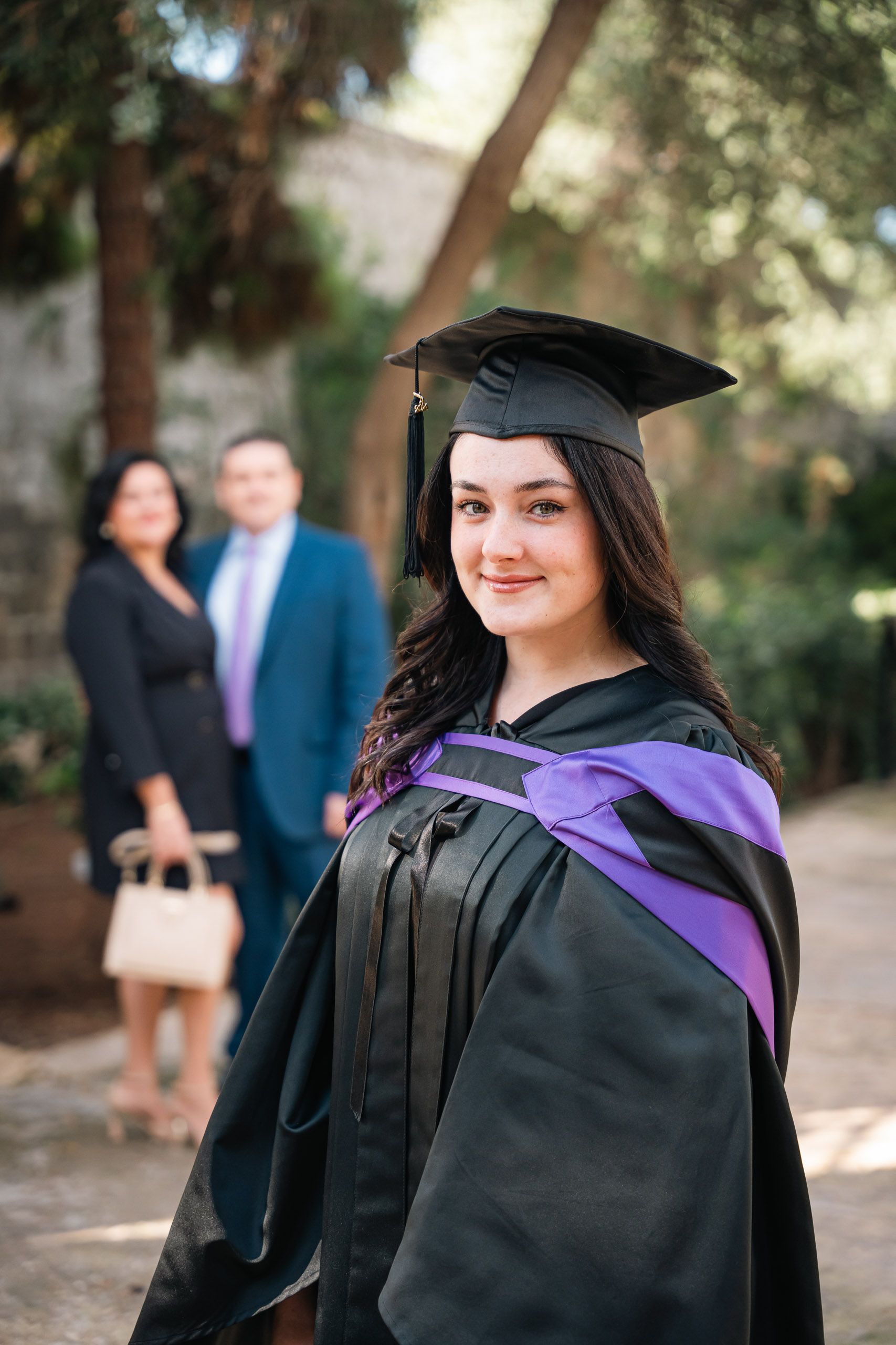 Graduate in a cap and purple-trimmed gown with her parents behind her at Romeo Romano Gardens in Malta.