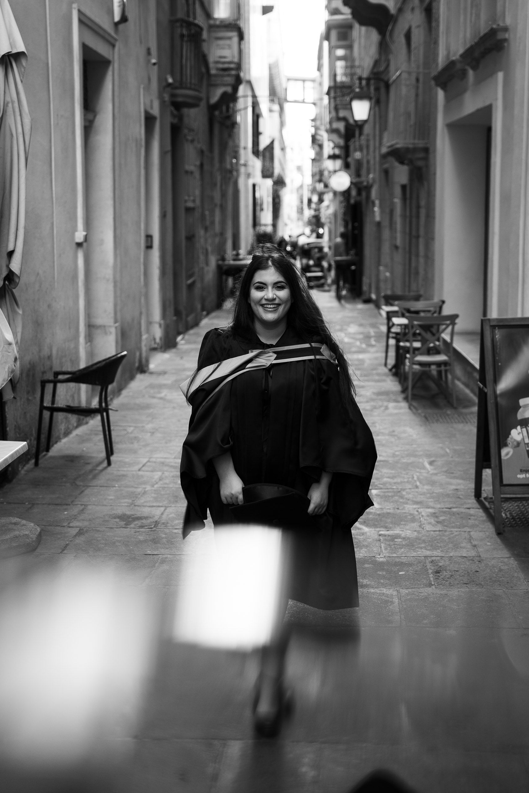 Black-and-white portrait of a smiling graduate holding her cap in a historic Valletta street.