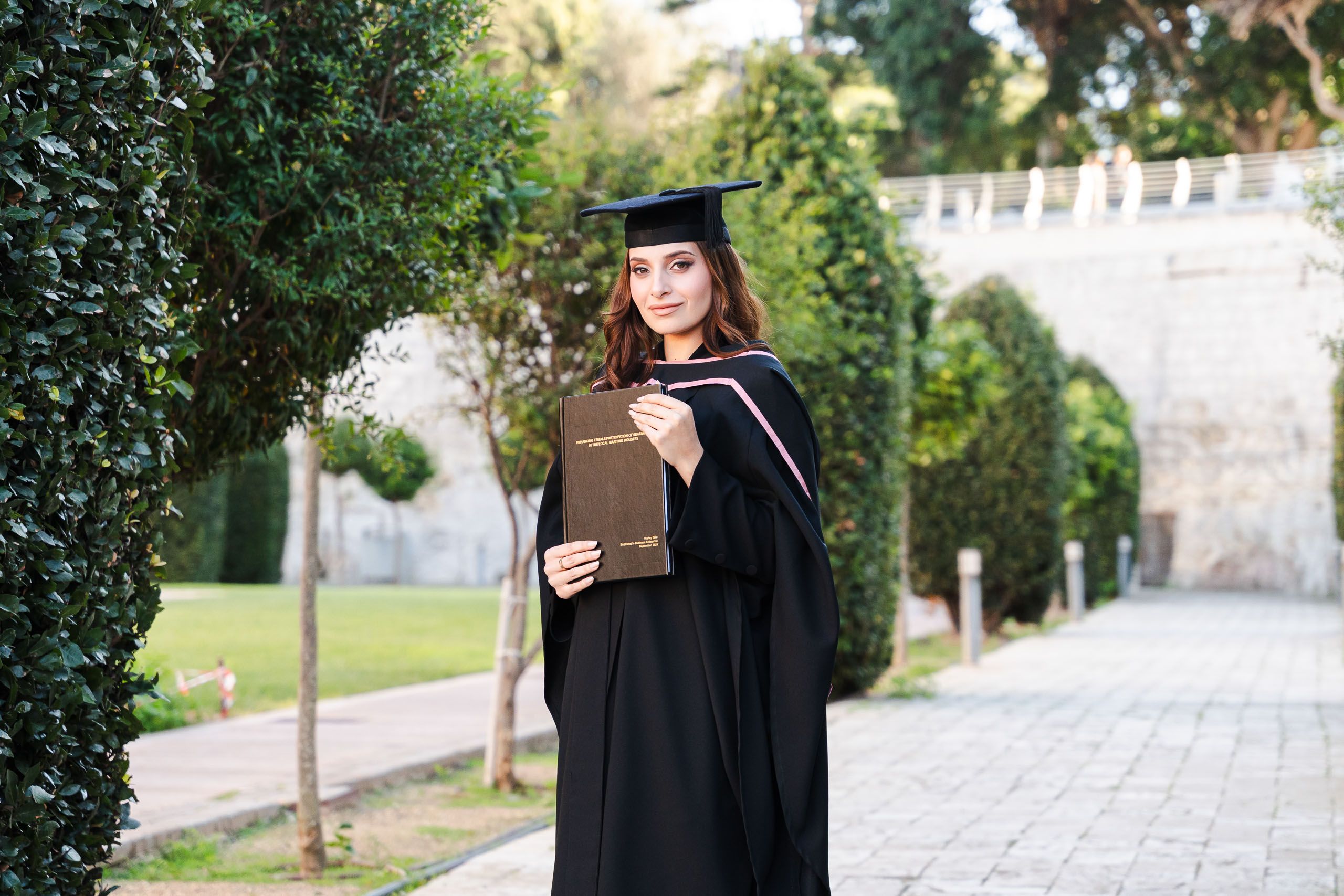 Graduate wearing a cap and gown while holding her dissertation in the gardens outside Mdina, Malta.