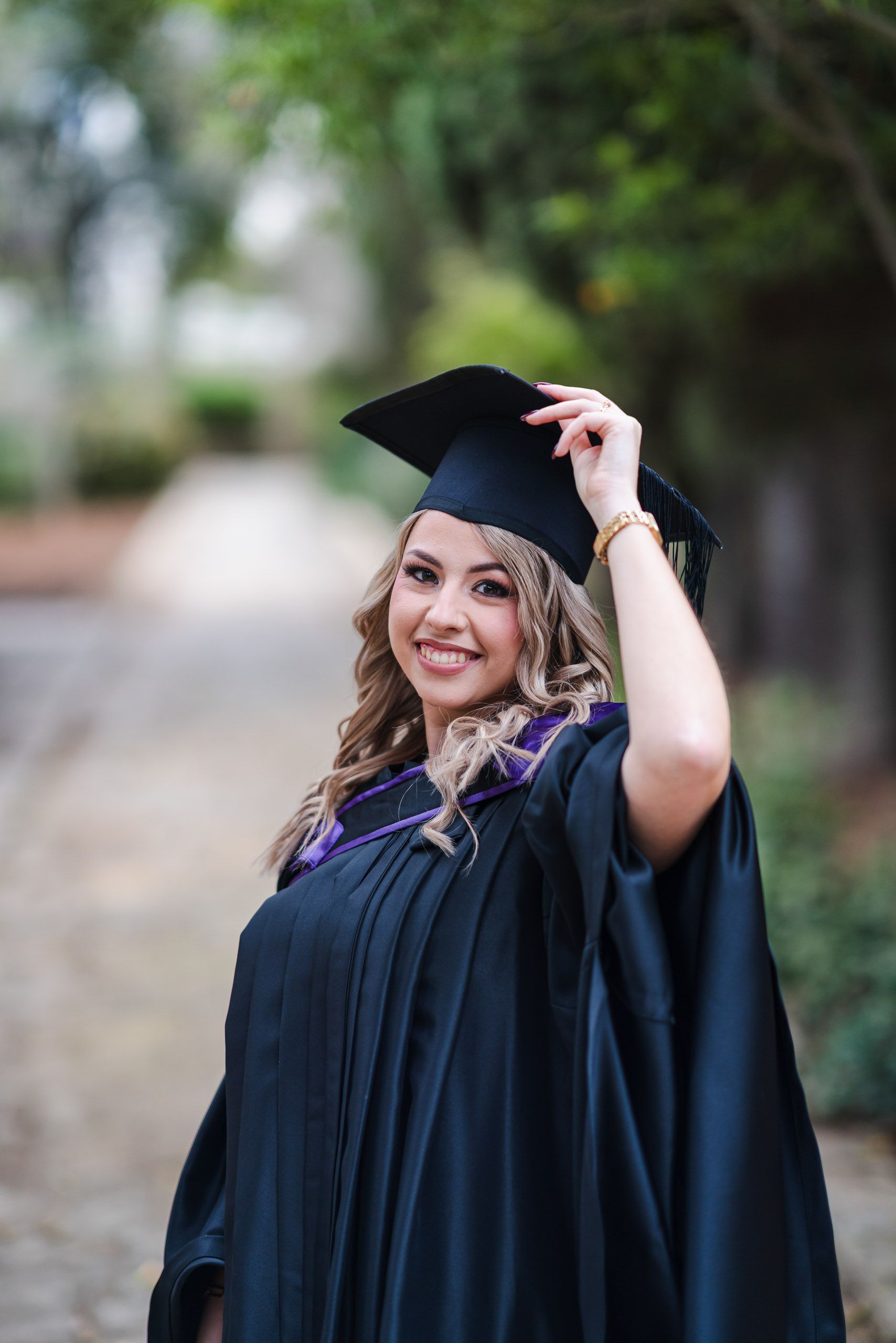 Smiling graduate adjusting her cap while wearing a purple-trimmed gown at San Anton Gardens in Malta.