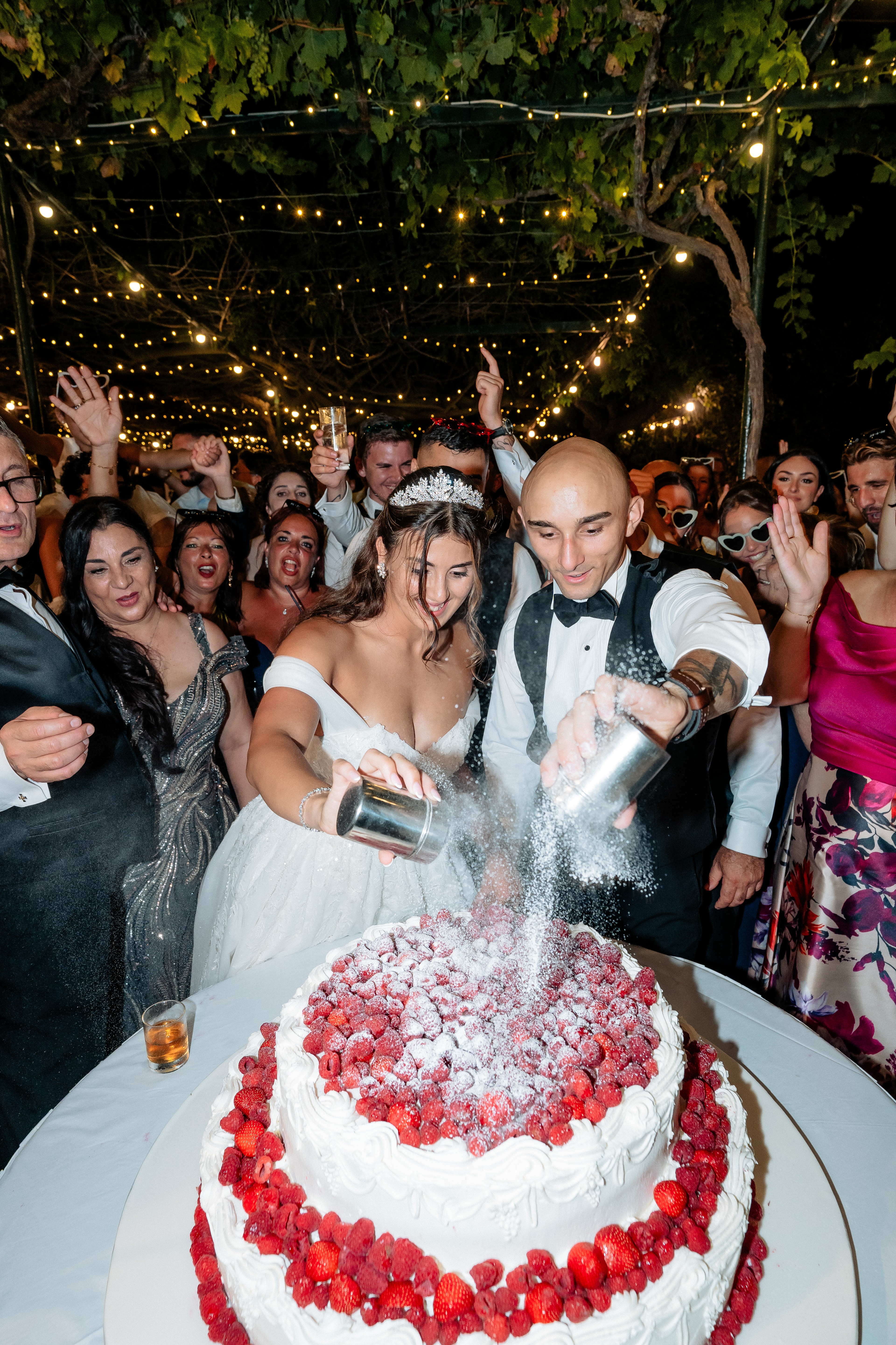 Bride and groom cutting their wedding cake at Razzett l-Abjad in Malta, surrounded by celebrating guests under string lights, photographed by Malta wedding photographer Justin Galea.