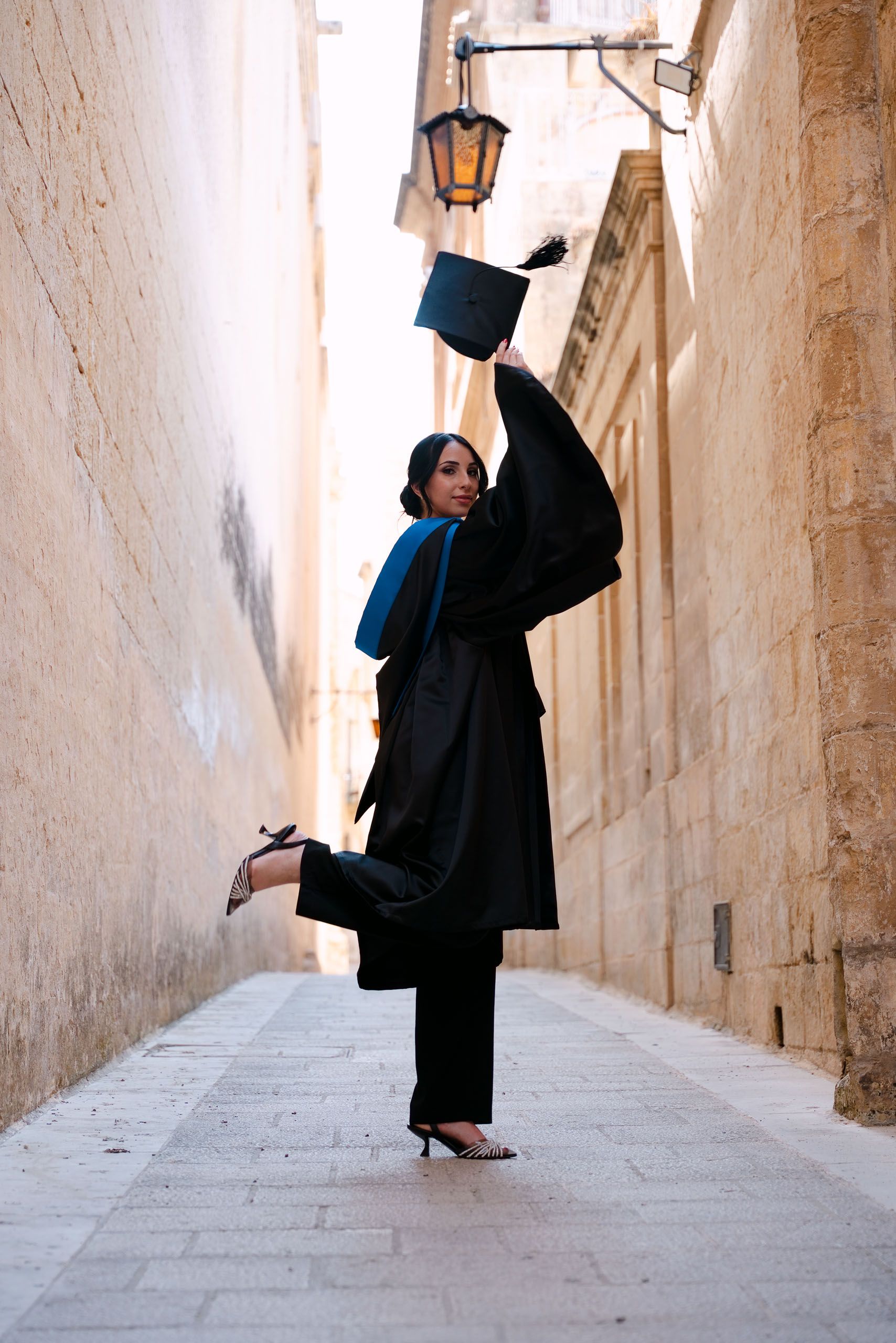 Graduate lifting her cap and posing playfully in one of the narrow historic streets of Mdina, Malta.