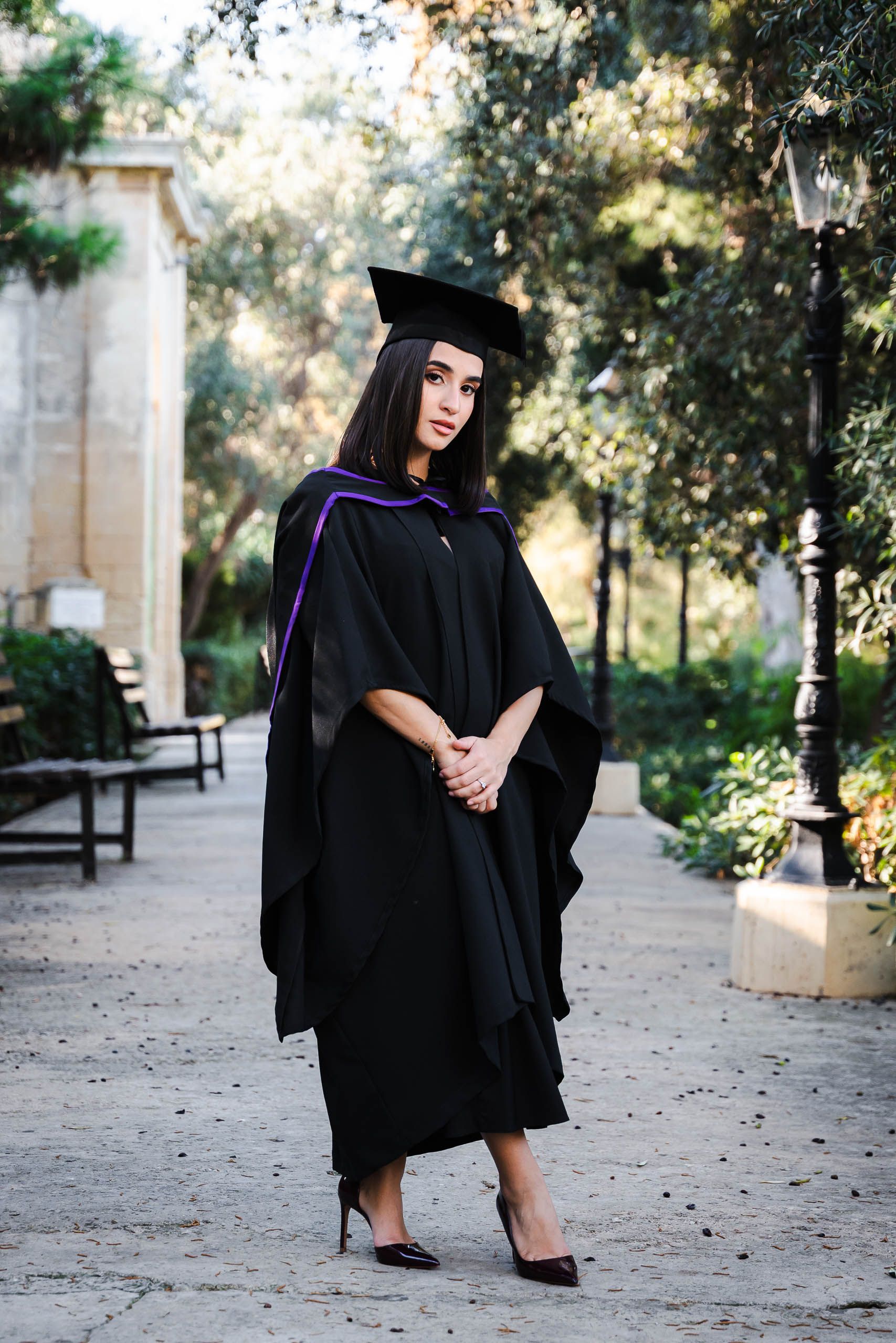 Full-length portrait of a graduate wearing a cap and purple-trimmed gown at Romeo Romano Gardens in Malta.