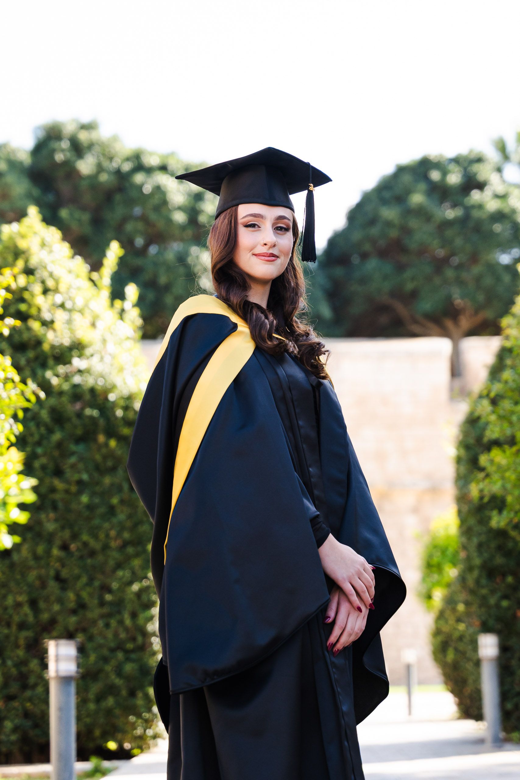 Female graduate wearing a cap and yellow-trimmed gown posing among the greenery at Mdina Ditch Gardens in Malta.