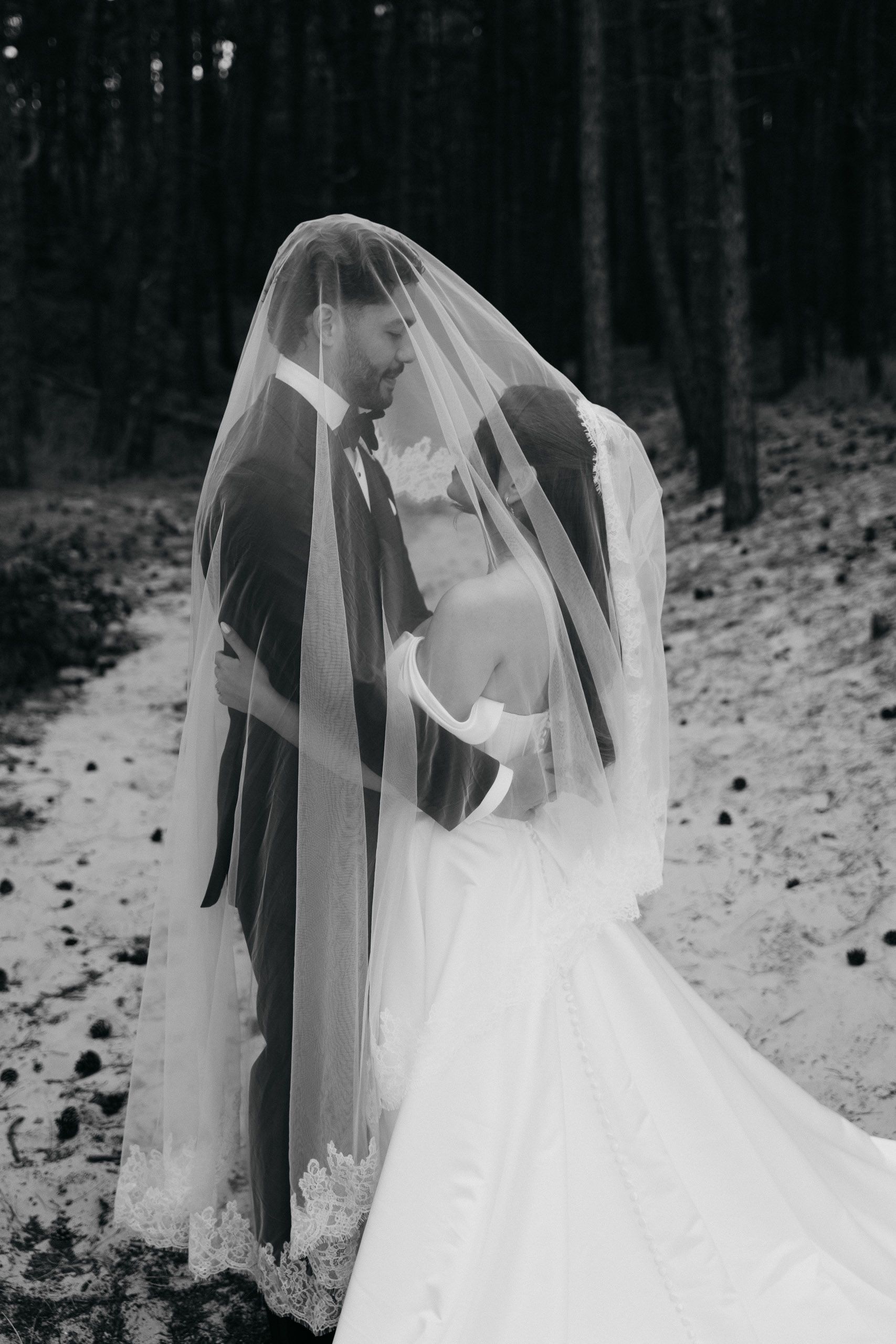 Bride and groom embracing beneath the wedding veil in the pine forest at Schoorlse Duinen, Netherlands.