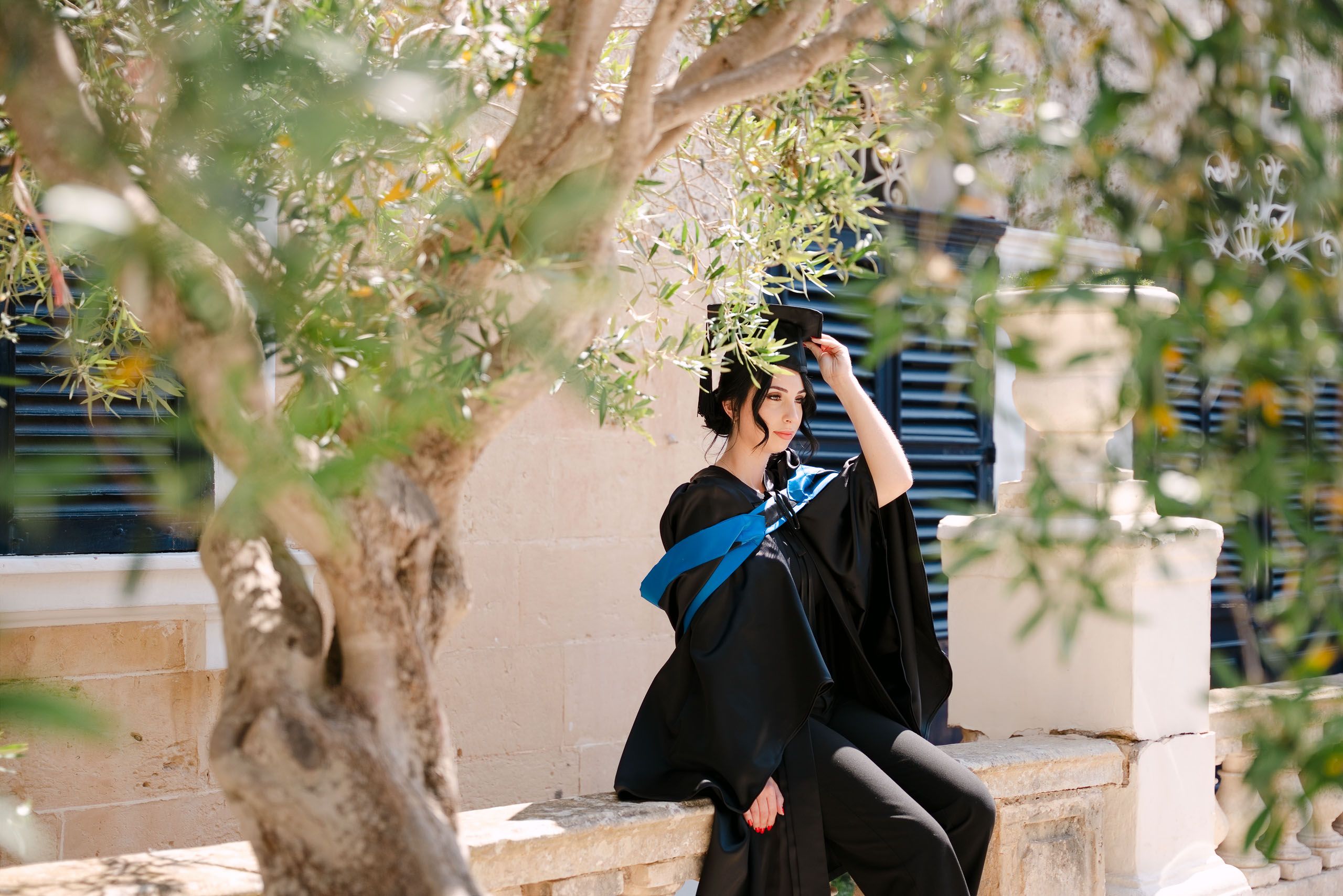 Graduate adjusting her cap while seated beneath the trees during a graduation photoshoot in Mdina and Rabat.