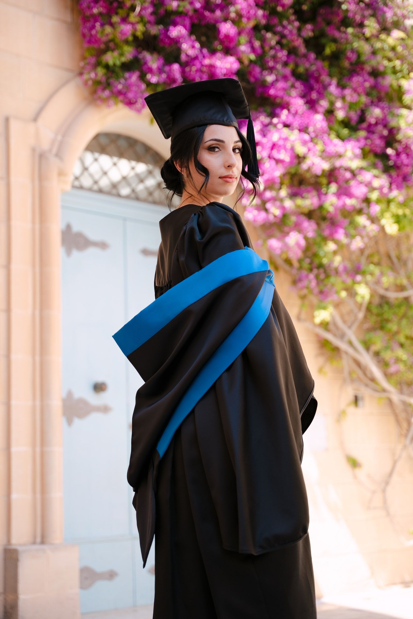 Female graduate in a blue-trimmed gown looking over her shoulder beside pink bougainvillea in Mdina, Malta.