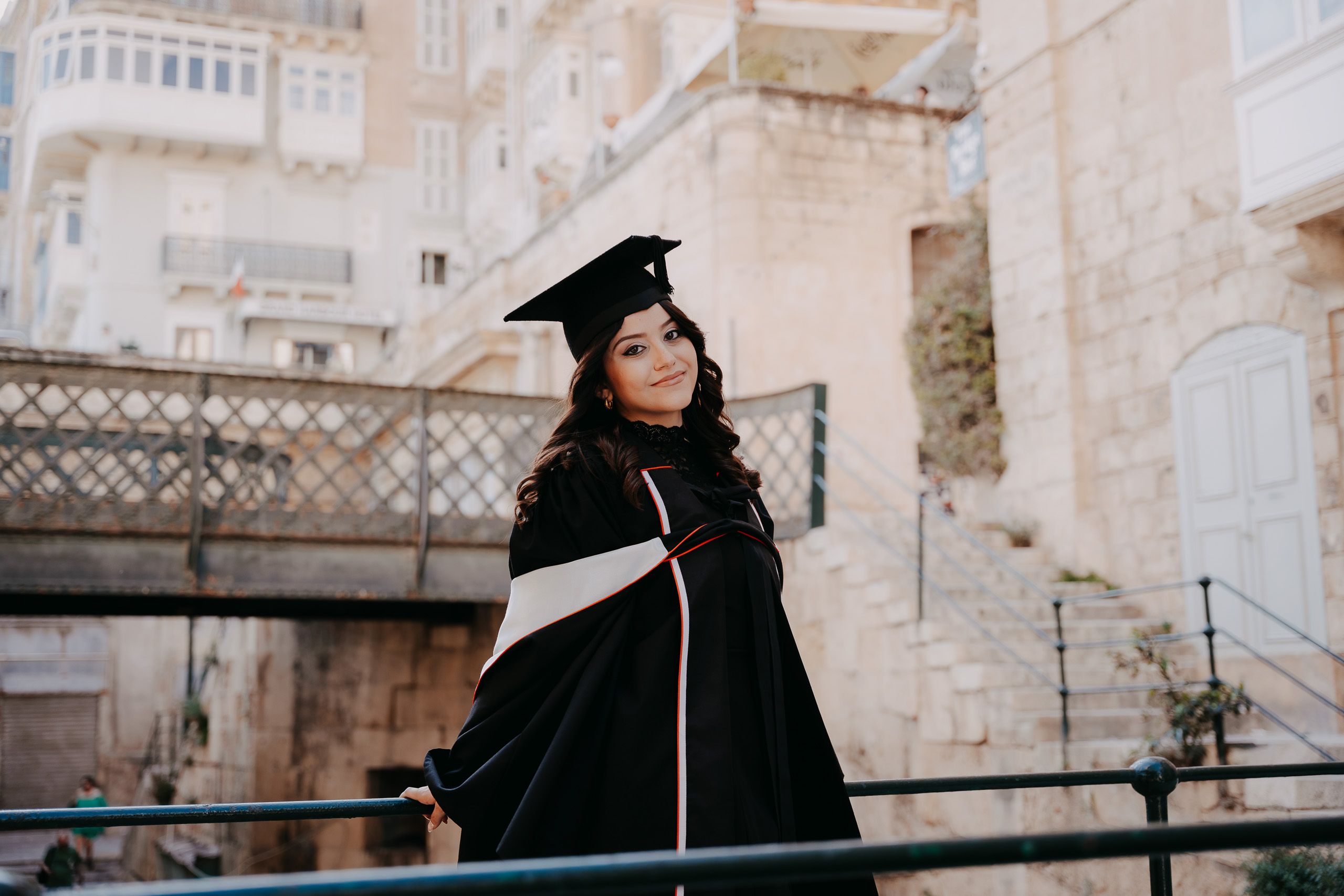 Graduate wearing a cap and gown posing on the Victoria Gate Bridge in Valletta, Malta.