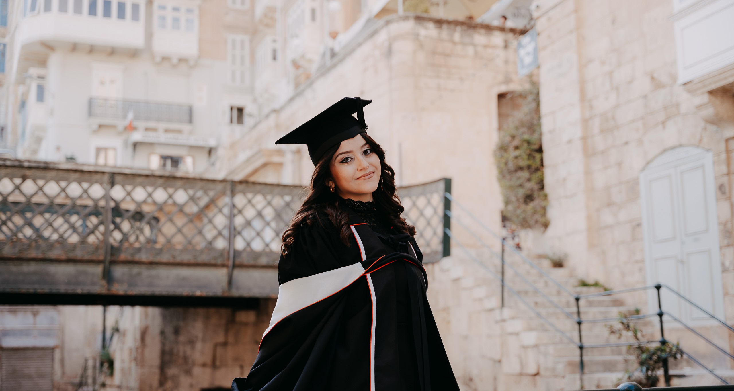 Graduate wearing a cap and gown posing on the Victoria Gate Bridge in Valletta, Malta.