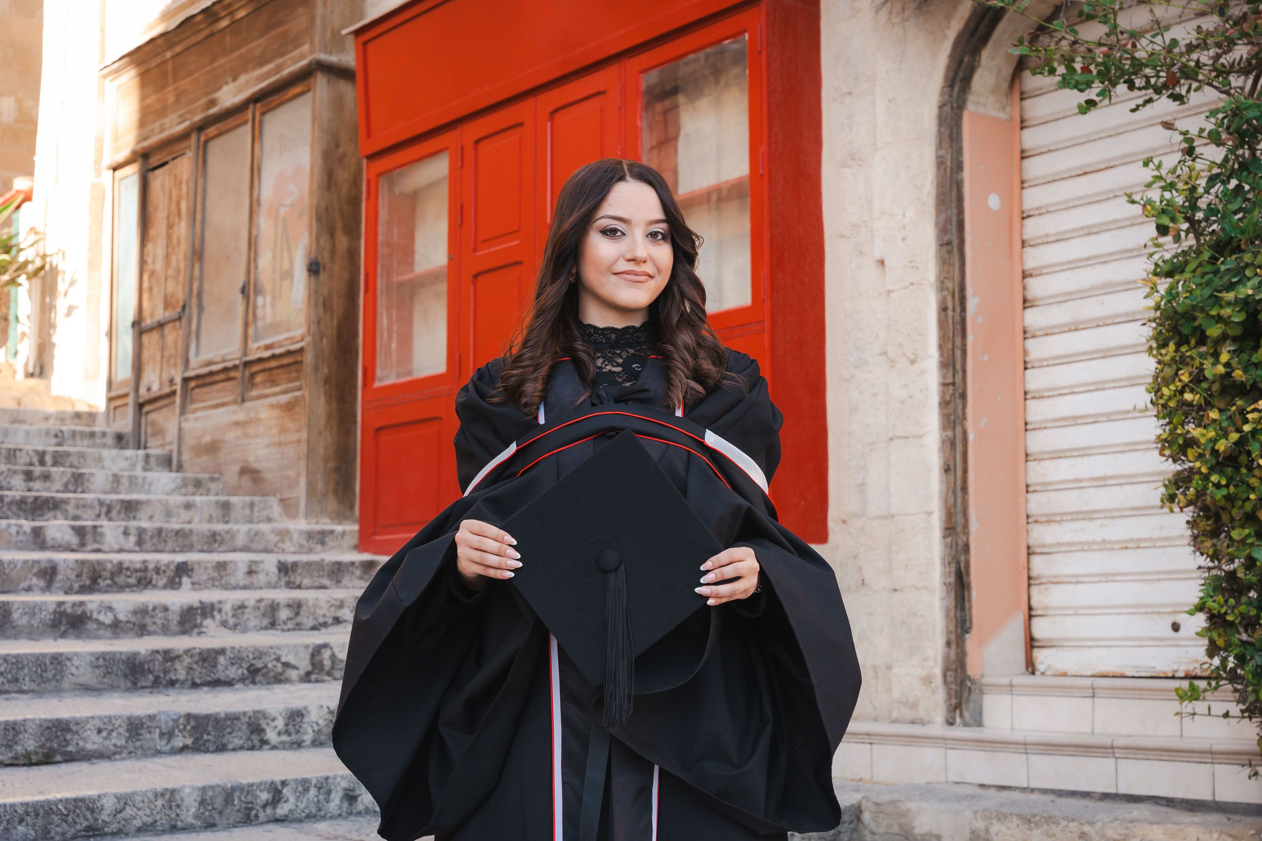Female graduate holding her cap while posing beside red doors and stone steps near Victoria Gate in Valletta.