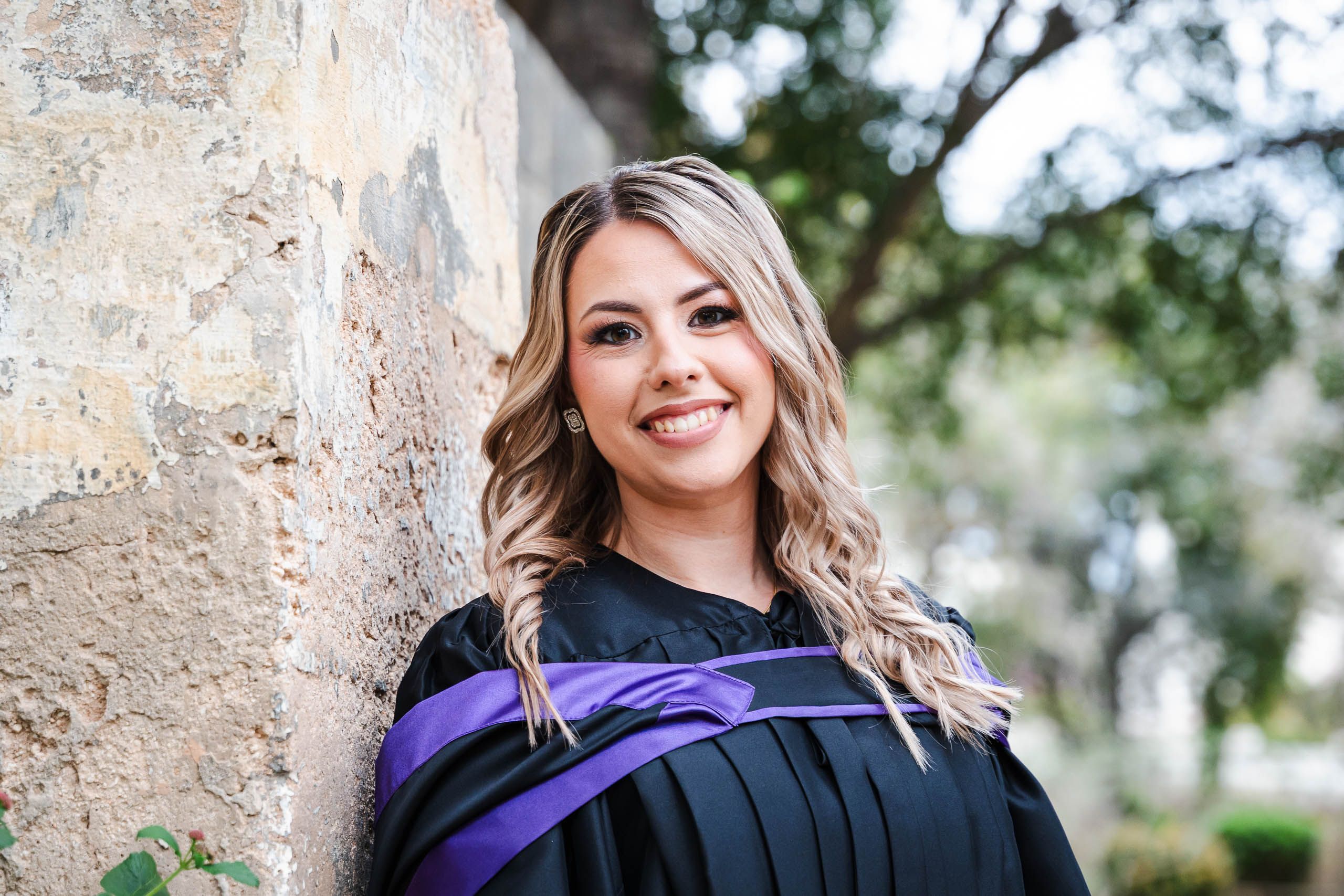 Graduate wearing a purple-trimmed gown leaning against a stone wall at San Anton Gardens in Malta.
