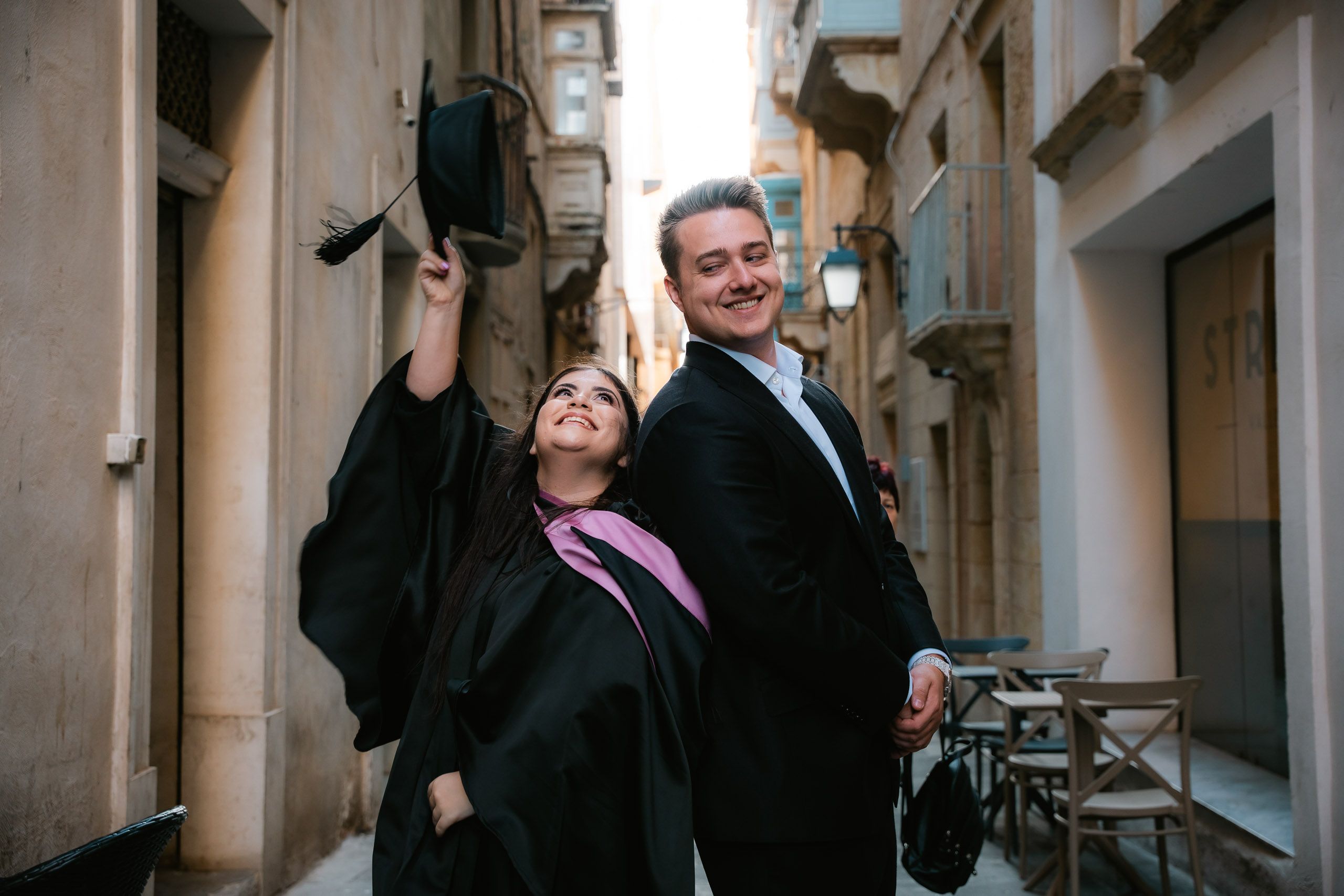Graduate celebrating with a companion while lifting her cap in a narrow street in Valletta, Malta.