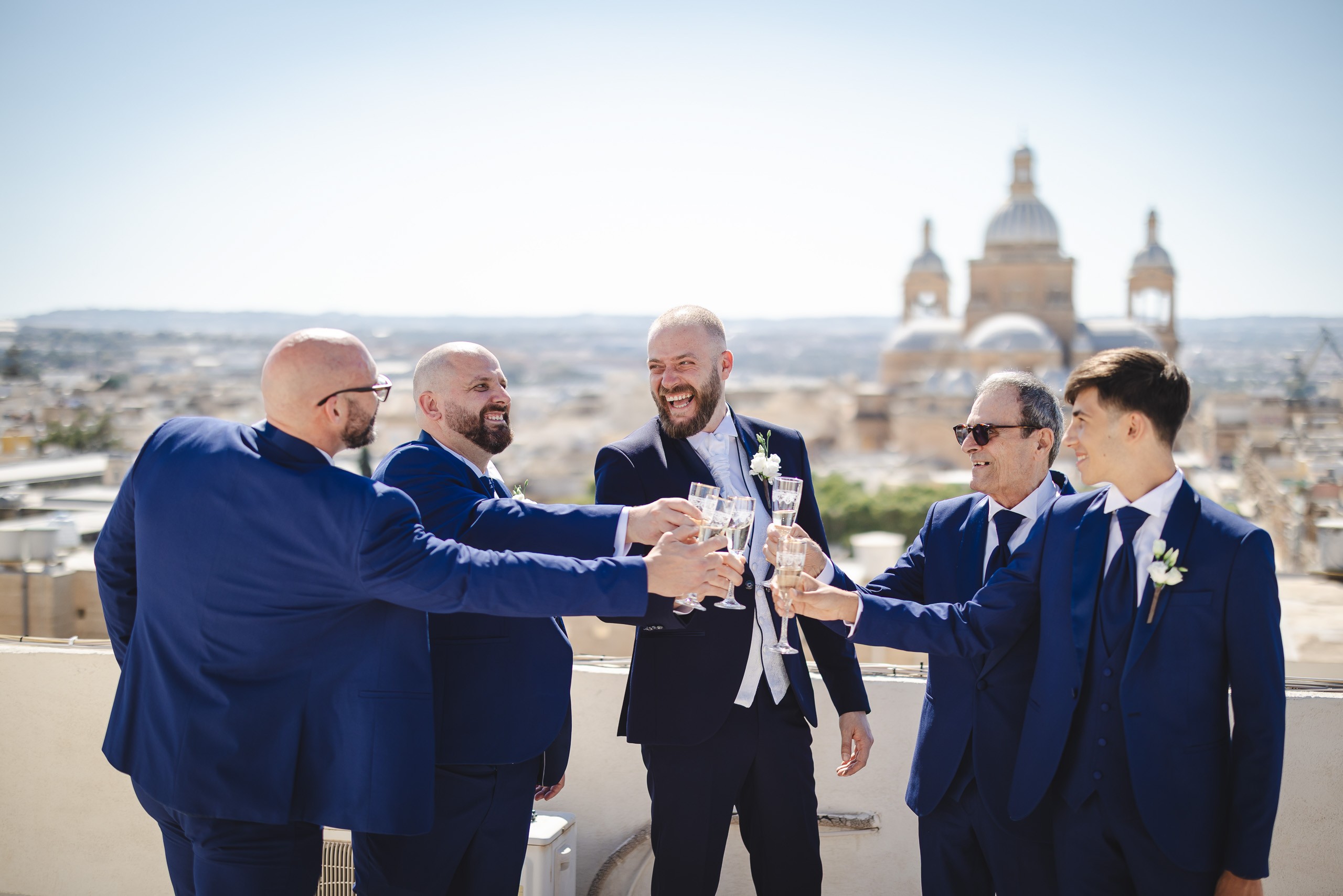 Groom and groomsmen raising champagne glasses on a rooftop with a Maltese church in the background.