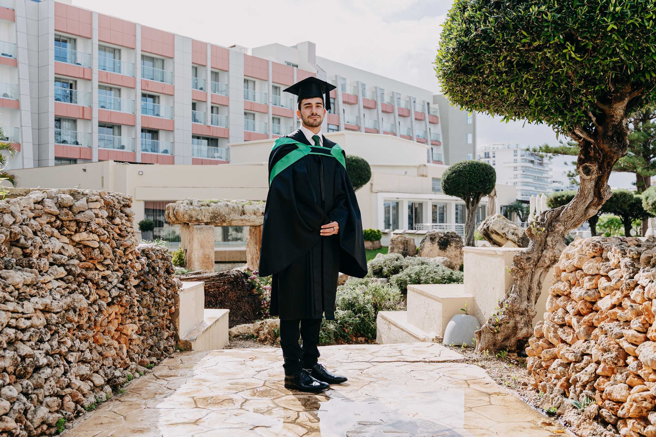 Full-length portrait of a male graduate in the gardens at Hilton Malta, surrounded by stone features and trees.
