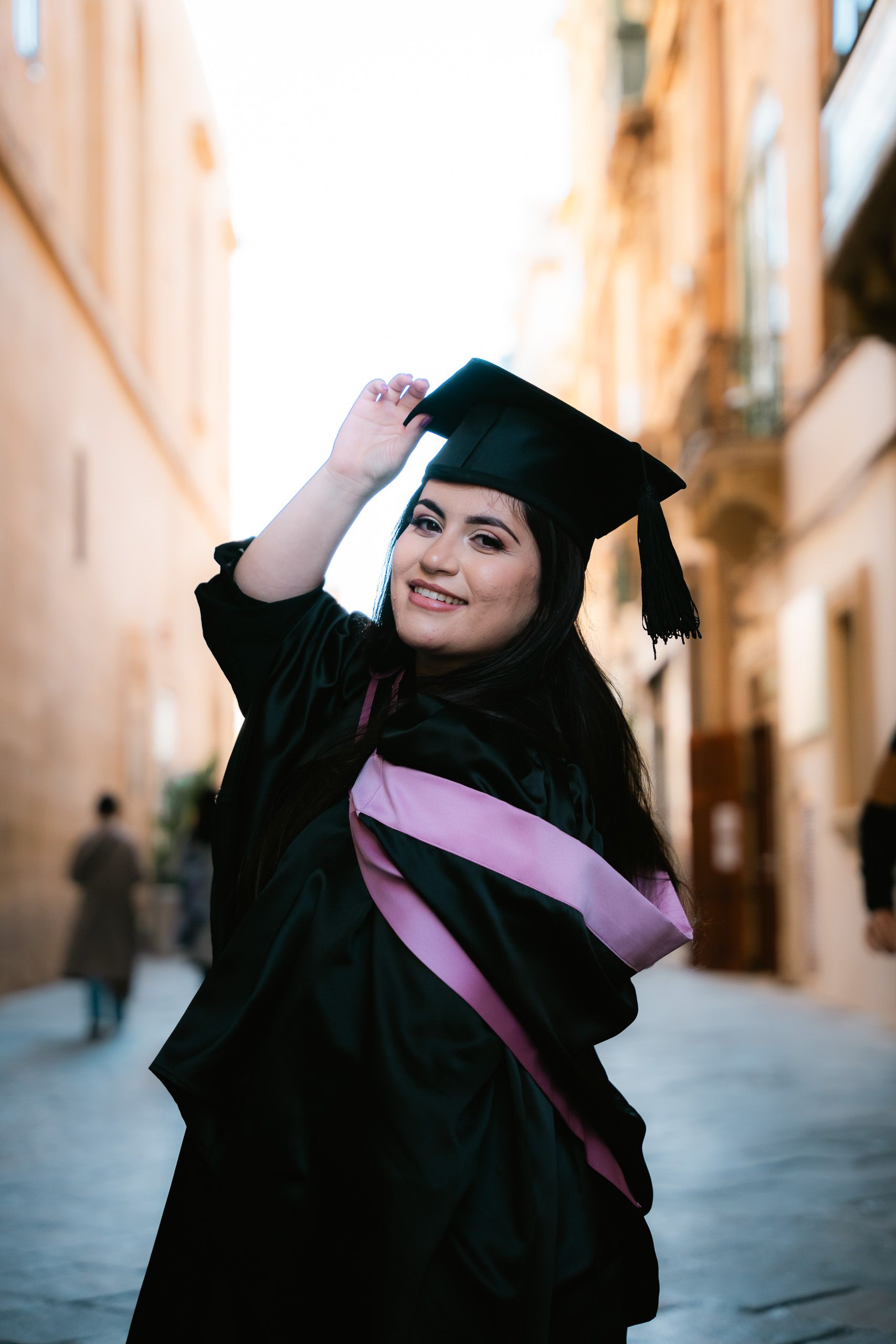Close-up portrait of a smiling graduate adjusting her cap in a sunlit Valletta street.