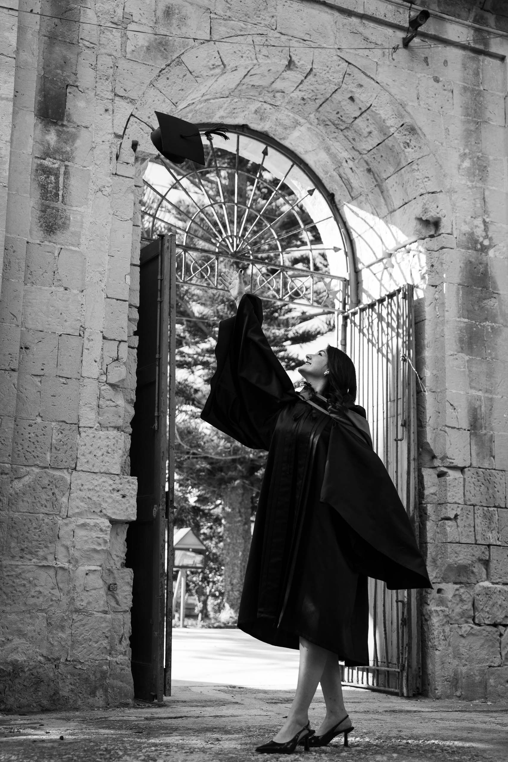 Black-and-white portrait of a graduate tossing her cap beneath the stone arch at Romeo Romano Gardens, Malta.