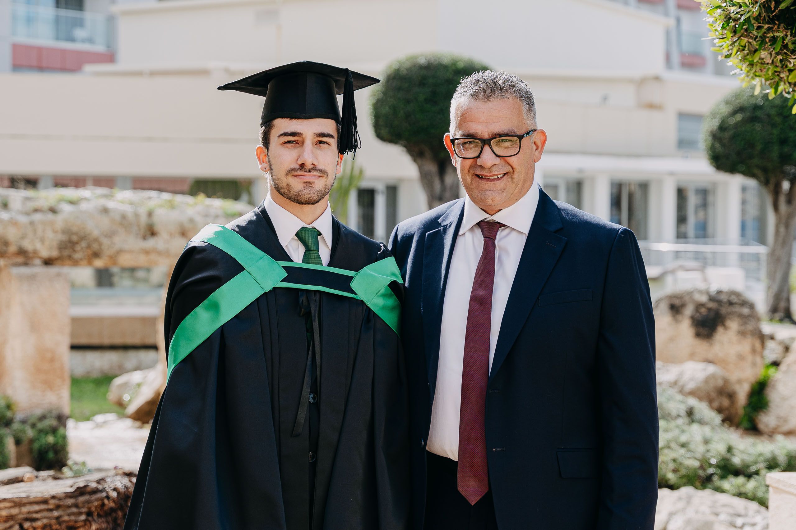 Male graduate in a green-trimmed gown posing with his father during a graduation photoshoot at Hilton Malta.