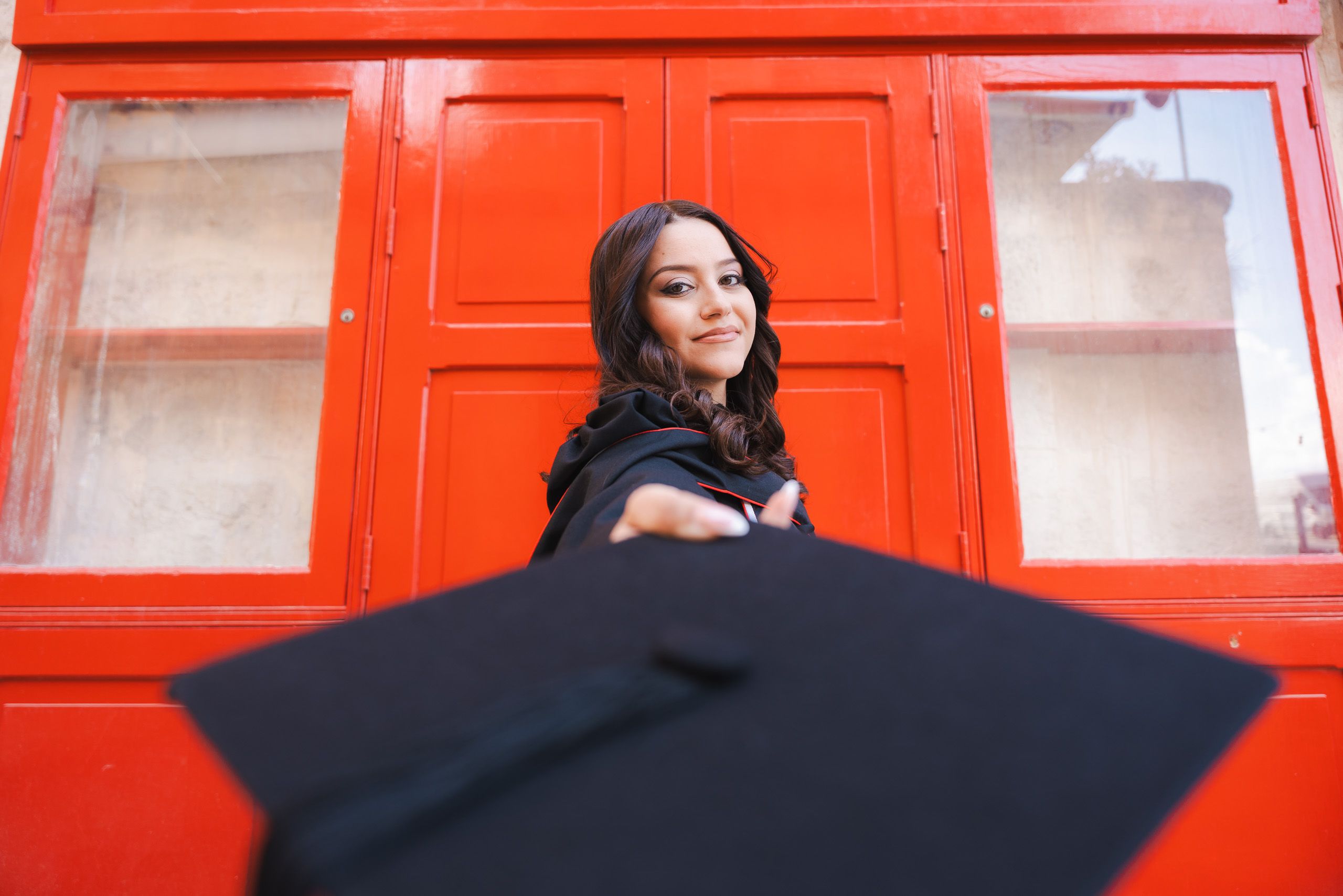 Female graduate reaching her cap towards the camera in front of a bright red doorway near Victoria Gate in Valletta.