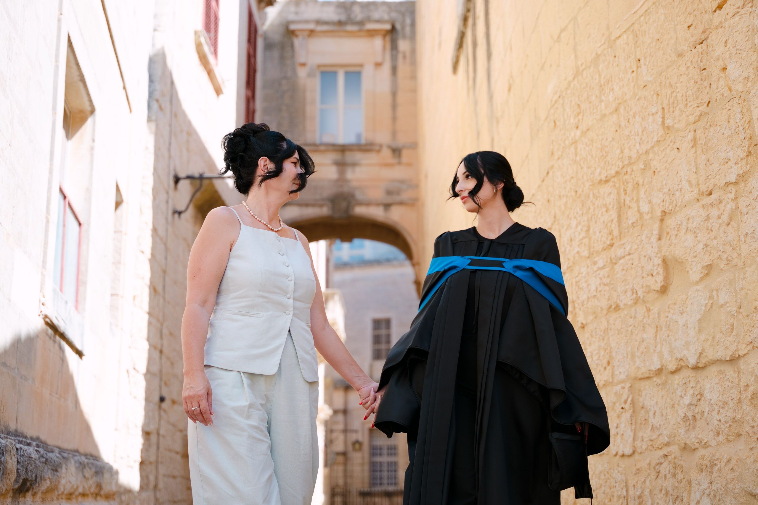 Female graduate holding hands with her mother while walking through a narrow limestone street in Mdina.