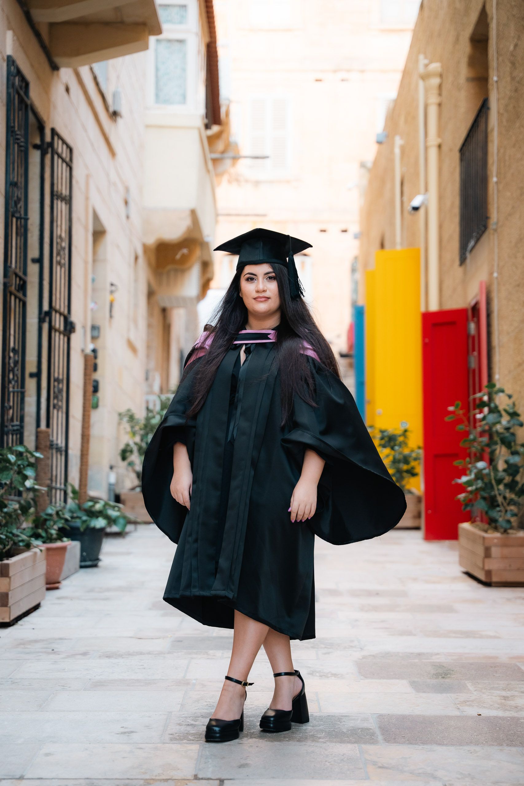 Female graduate in a cap and gown posing in a colourful narrow street in Valletta, Malta.