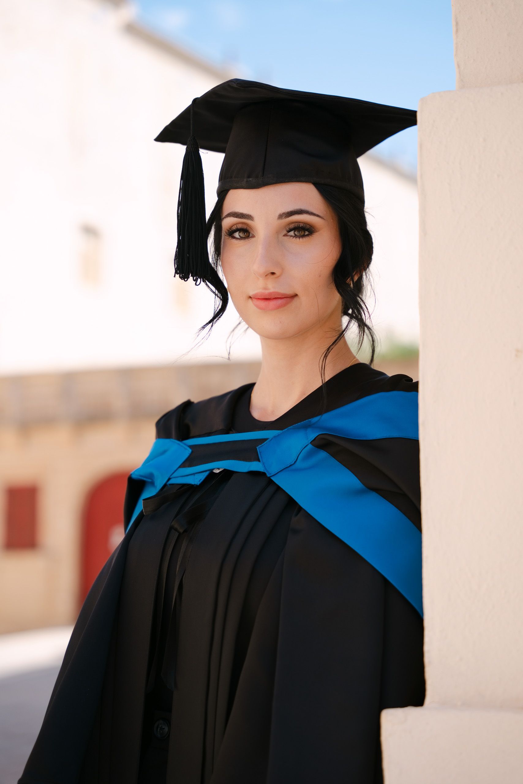 Female graduate in a blue-trimmed gown and cap leaning beside a stone column in Mdina, Malta.