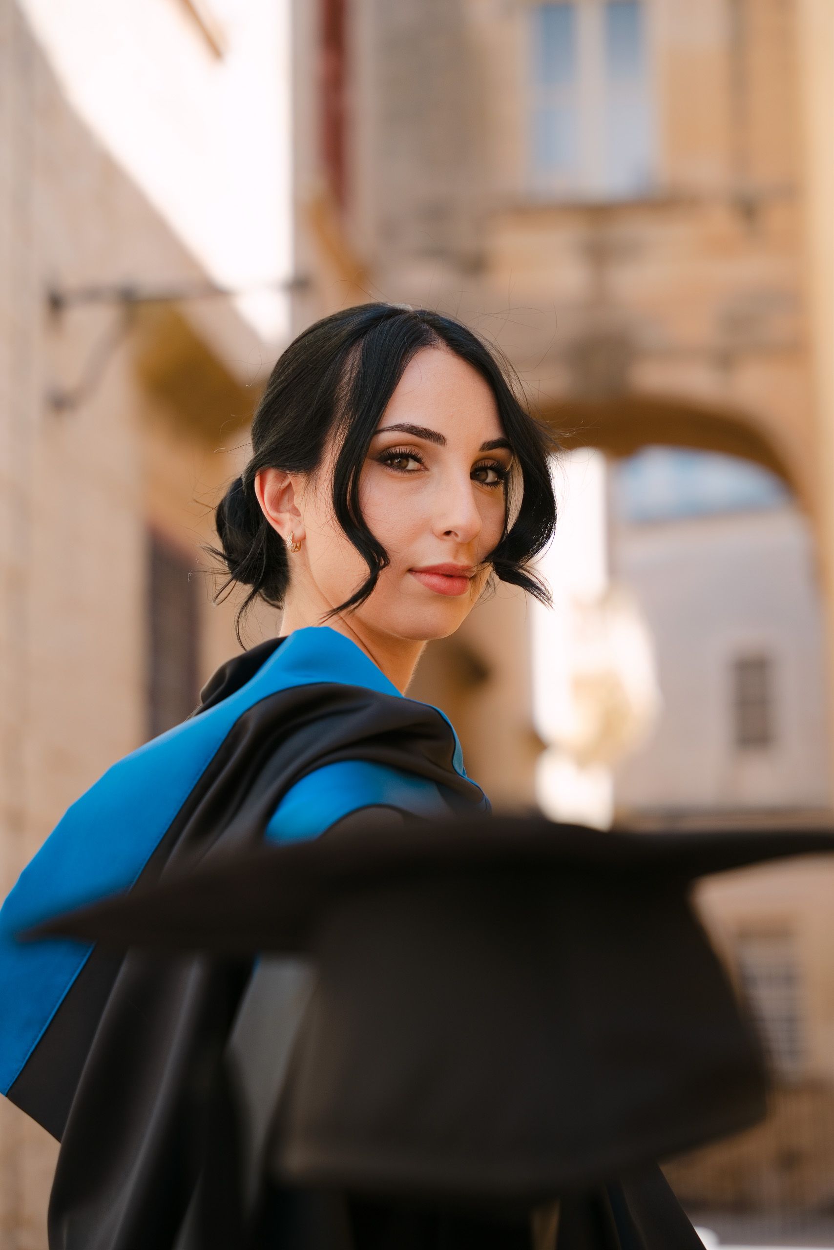 Close-up portrait of a graduate with her cap blurred in the foreground among the stone arches of Mdina.