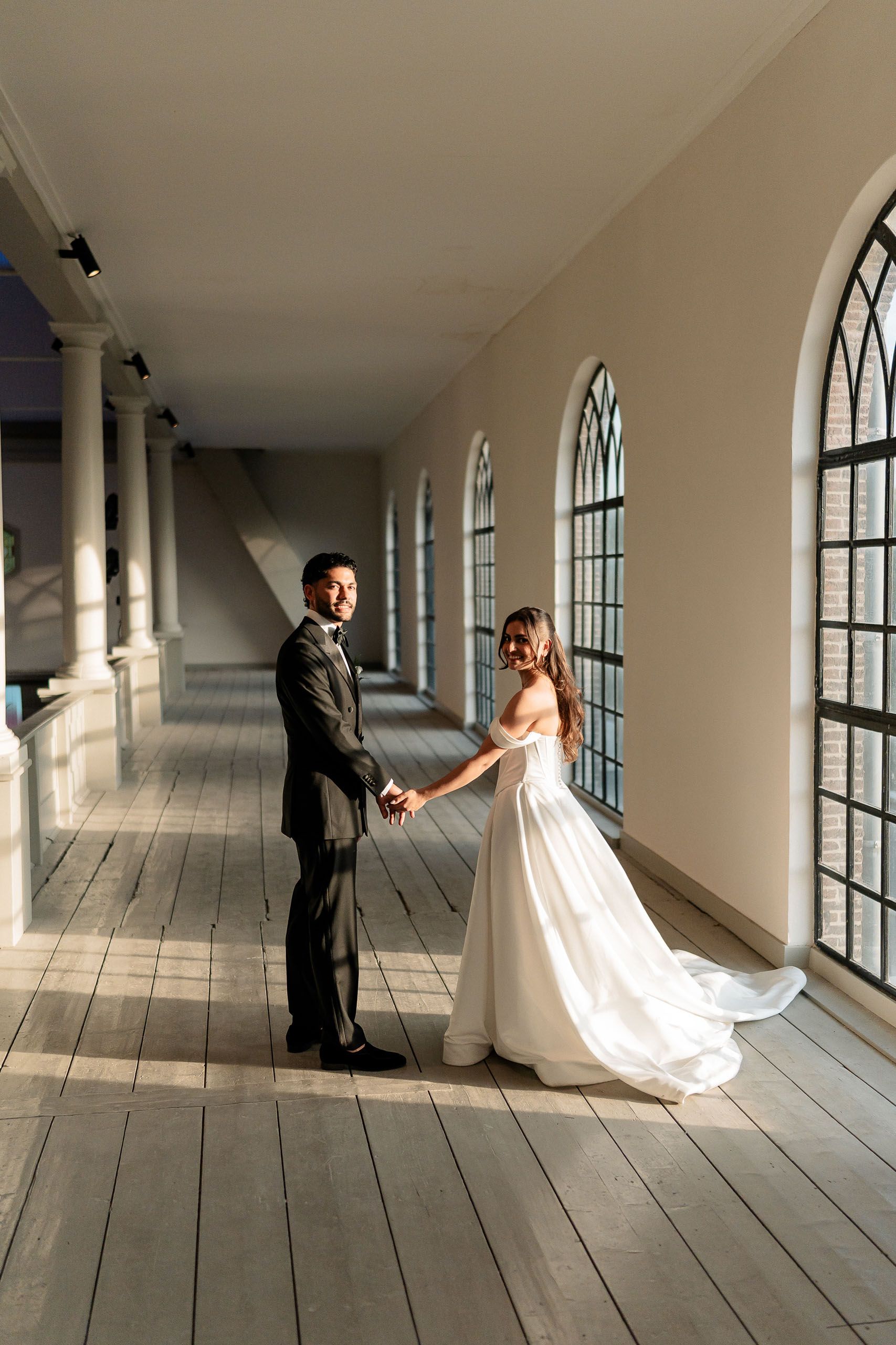 Bride and groom holding hands in the sunlit upper gallery of a church in Den Helder, Netherlands.