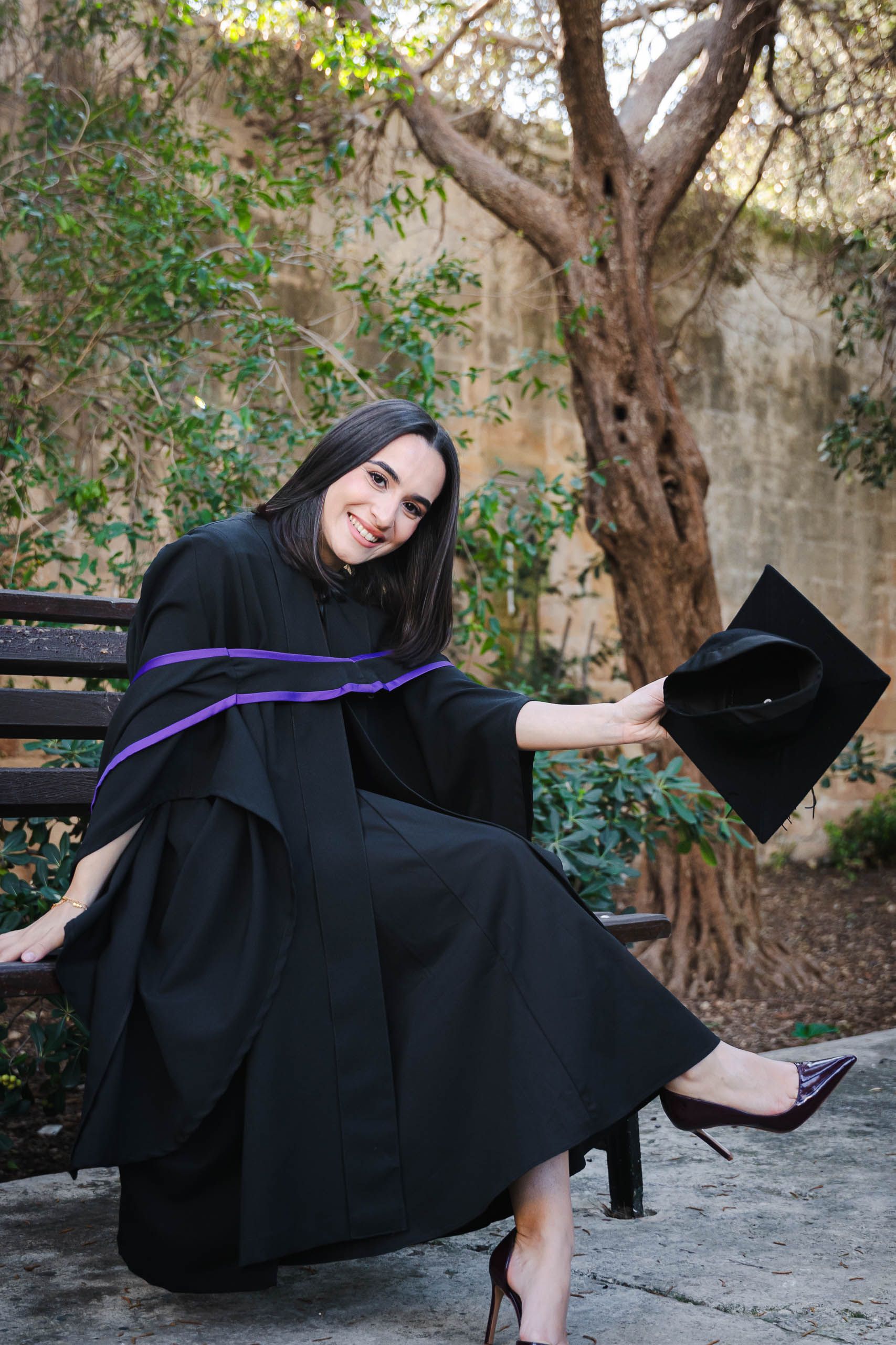 Smiling graduate seated on a bench and holding her cap at Romeo Romano Gardens in Malta.