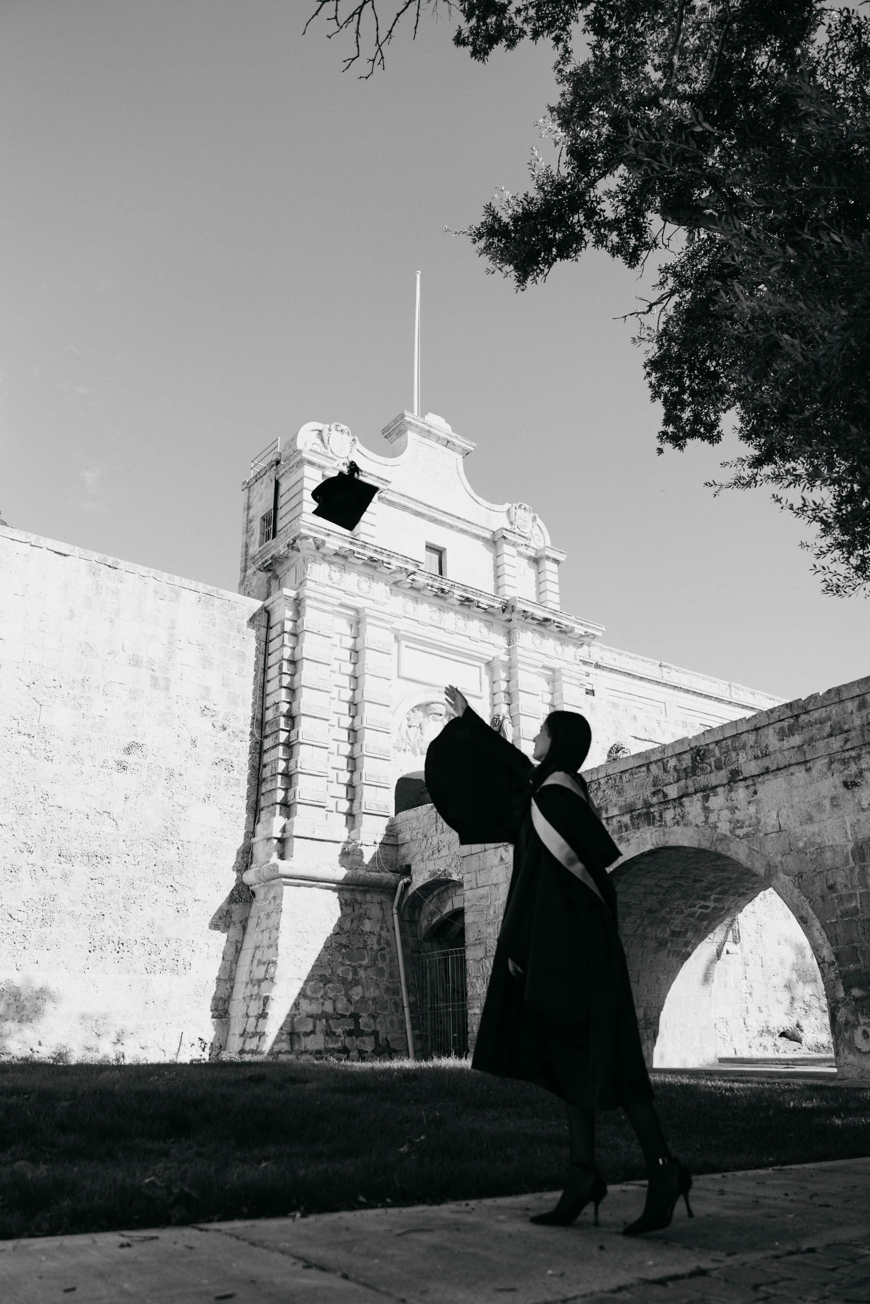 Black-and-white photograph of a graduate throwing her cap beneath the historic fortifications at Mdina Ditch Gardens.