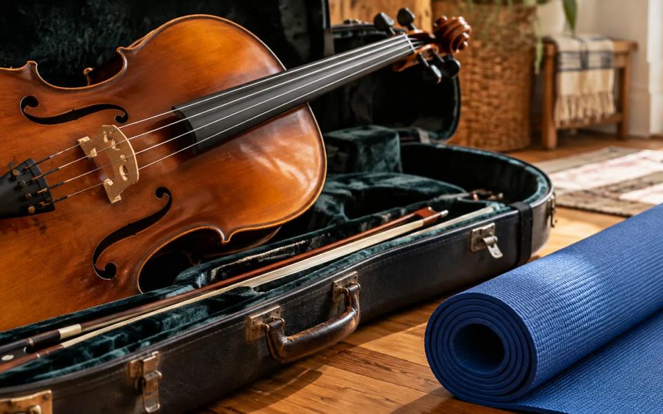 A cello in an open protective case beside a rolled blue mat on a wooden floor.