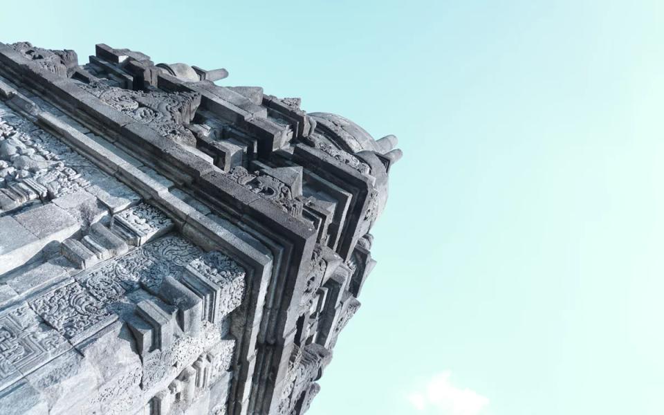 Upward view of carved stone architecture at Prambanan against a pale blue sky.