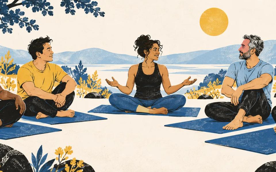 A group seated on blue mats outdoors, listening to a teacher with open hands.