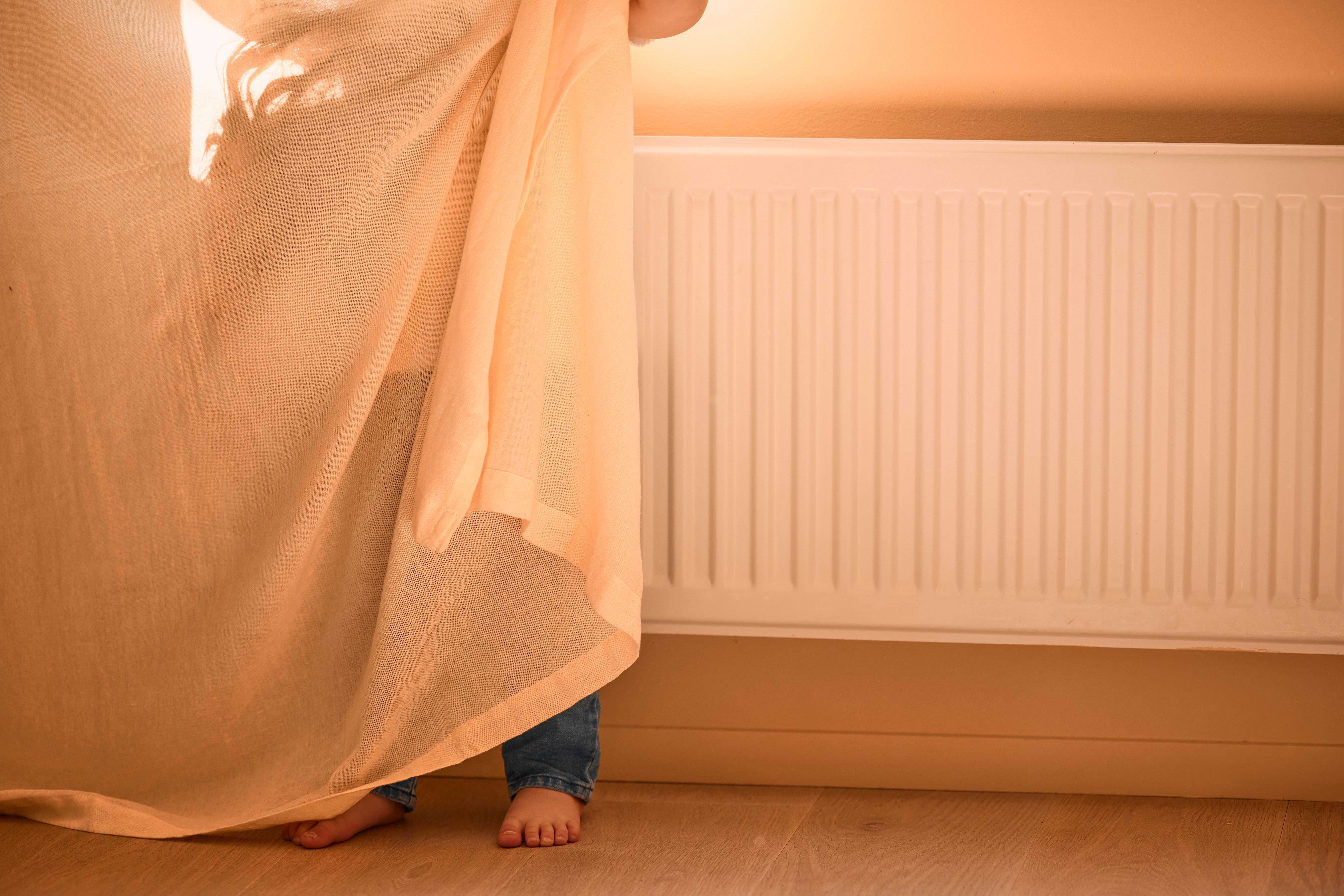 Barn i leilighet gjemmer seg bak en gardin. Bak barnet er det en radiator.