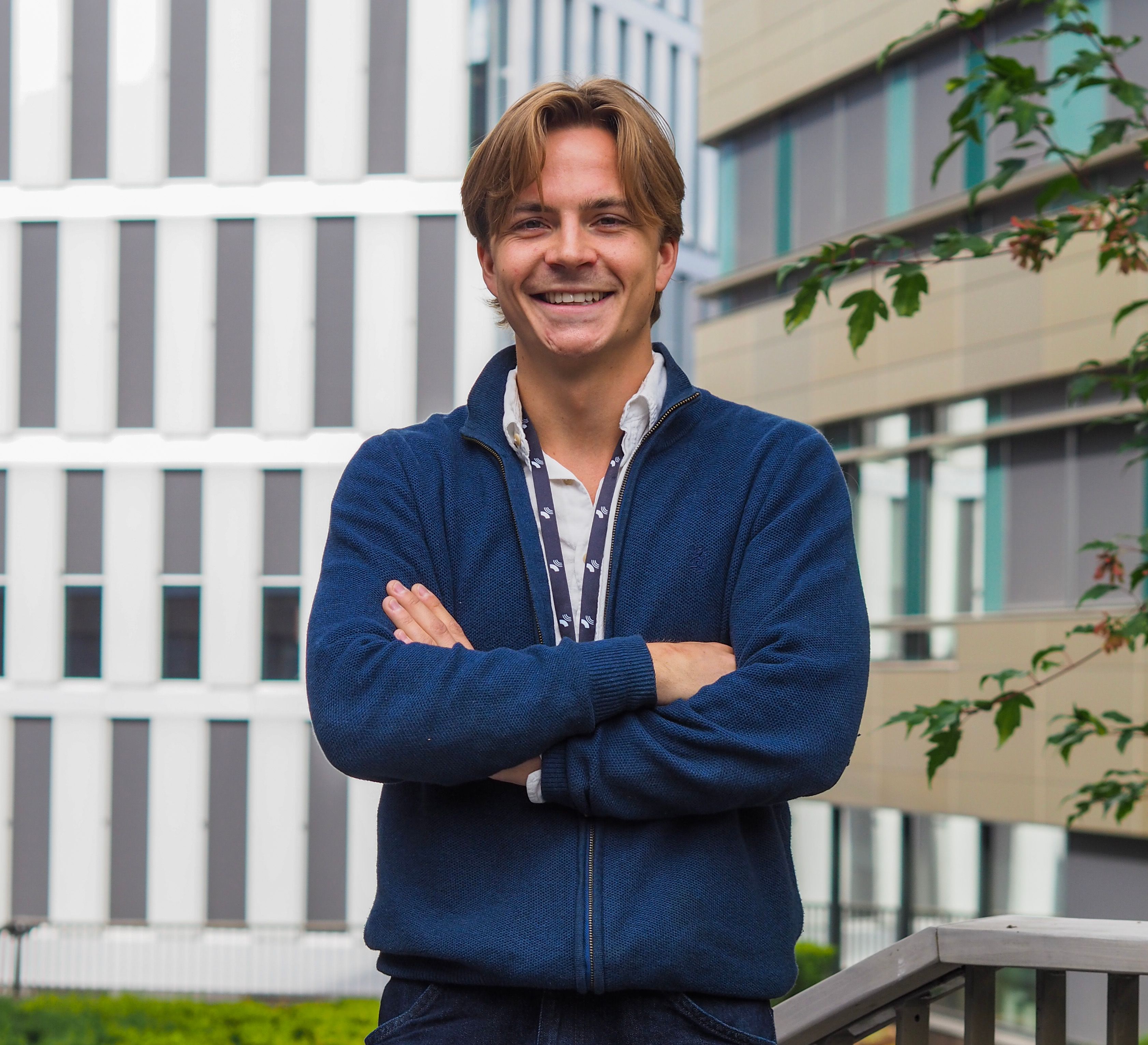 a man in a blue sweater is standing in front of a building with his arms crossed and smiling .