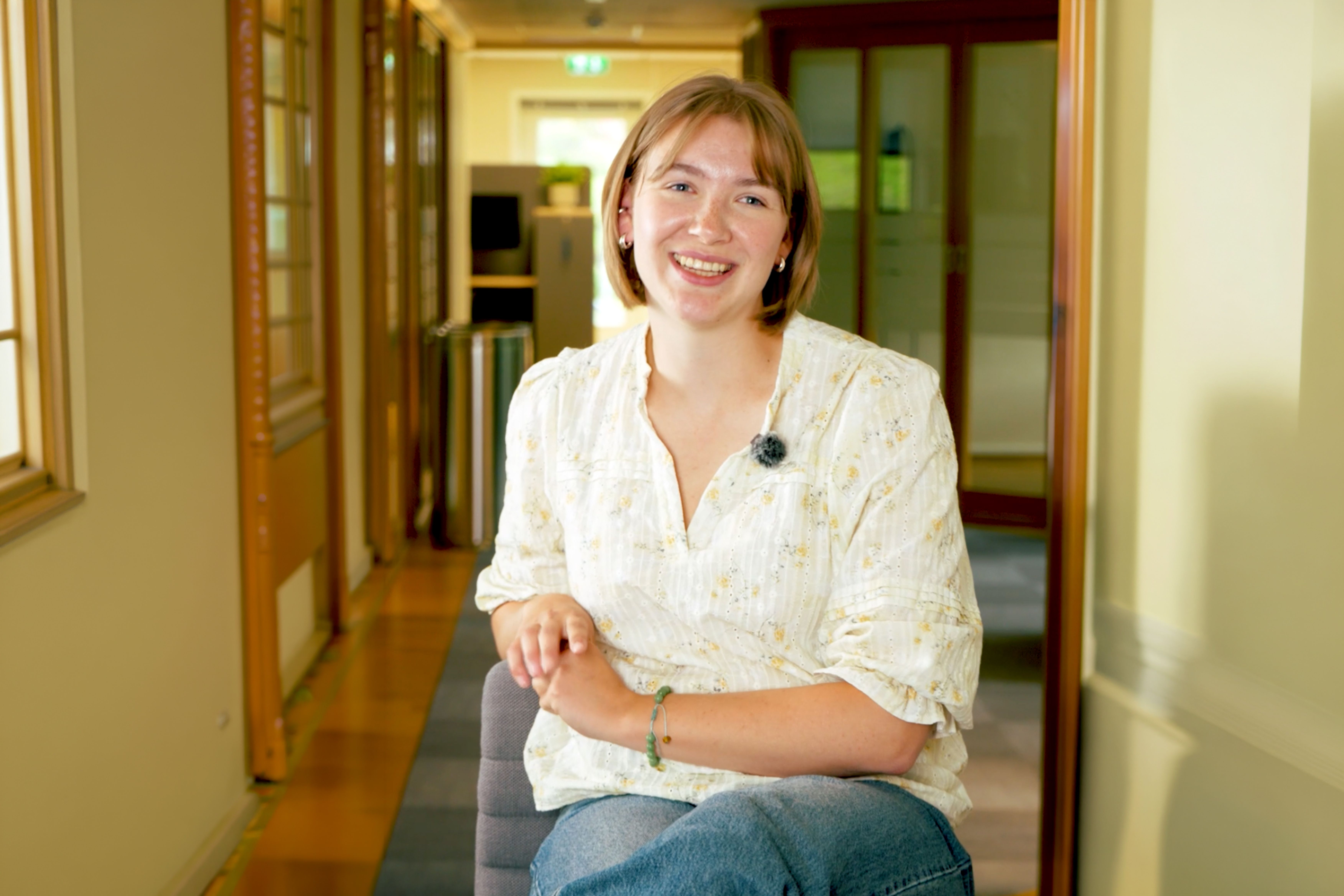 A smiling young woman with a mic clipped to her shirt sits in a hallway.