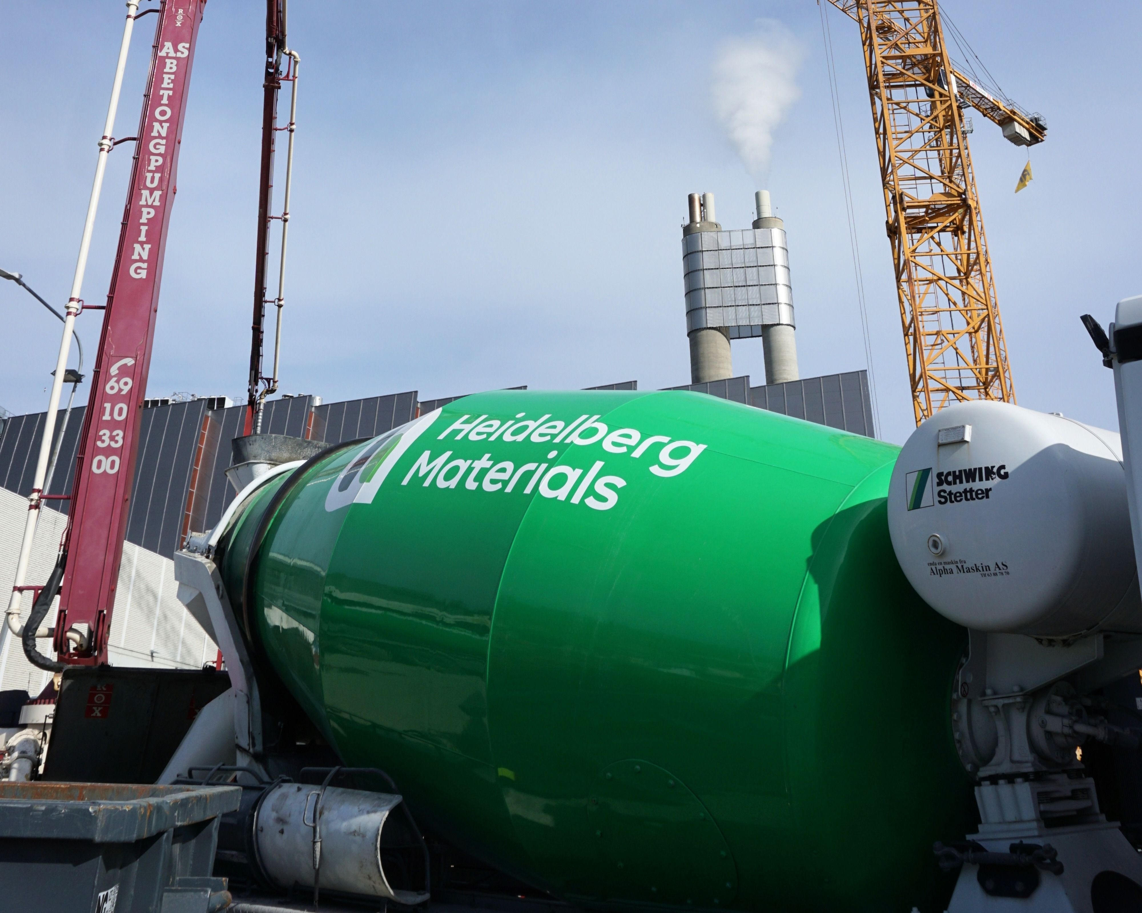 A green Heidelberg Materials cement mixer truck and a red concrete pump arm with an industrial background featuring smokestacks and a crane.