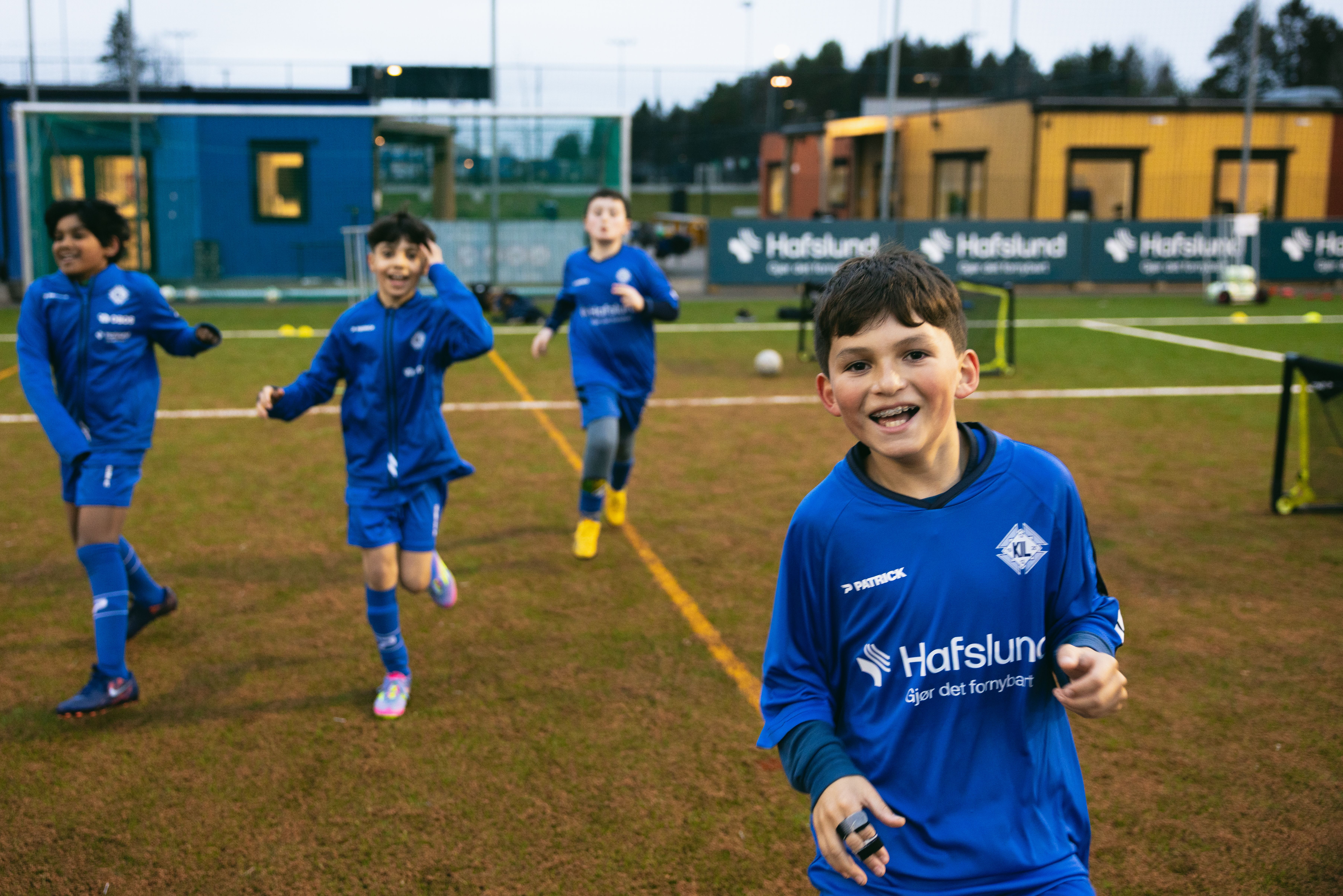 En smilende gutt i en blå fotballdrakt står på en kunstgressbane, med tre andre gutter som løper bak ham.