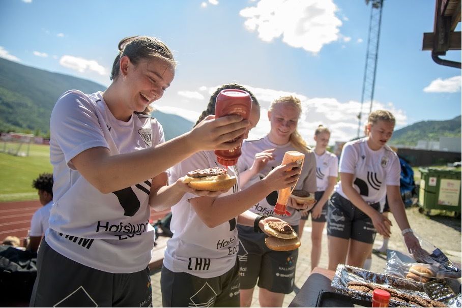 Fotballjenter spiser grillet hamburger ute i solen. 