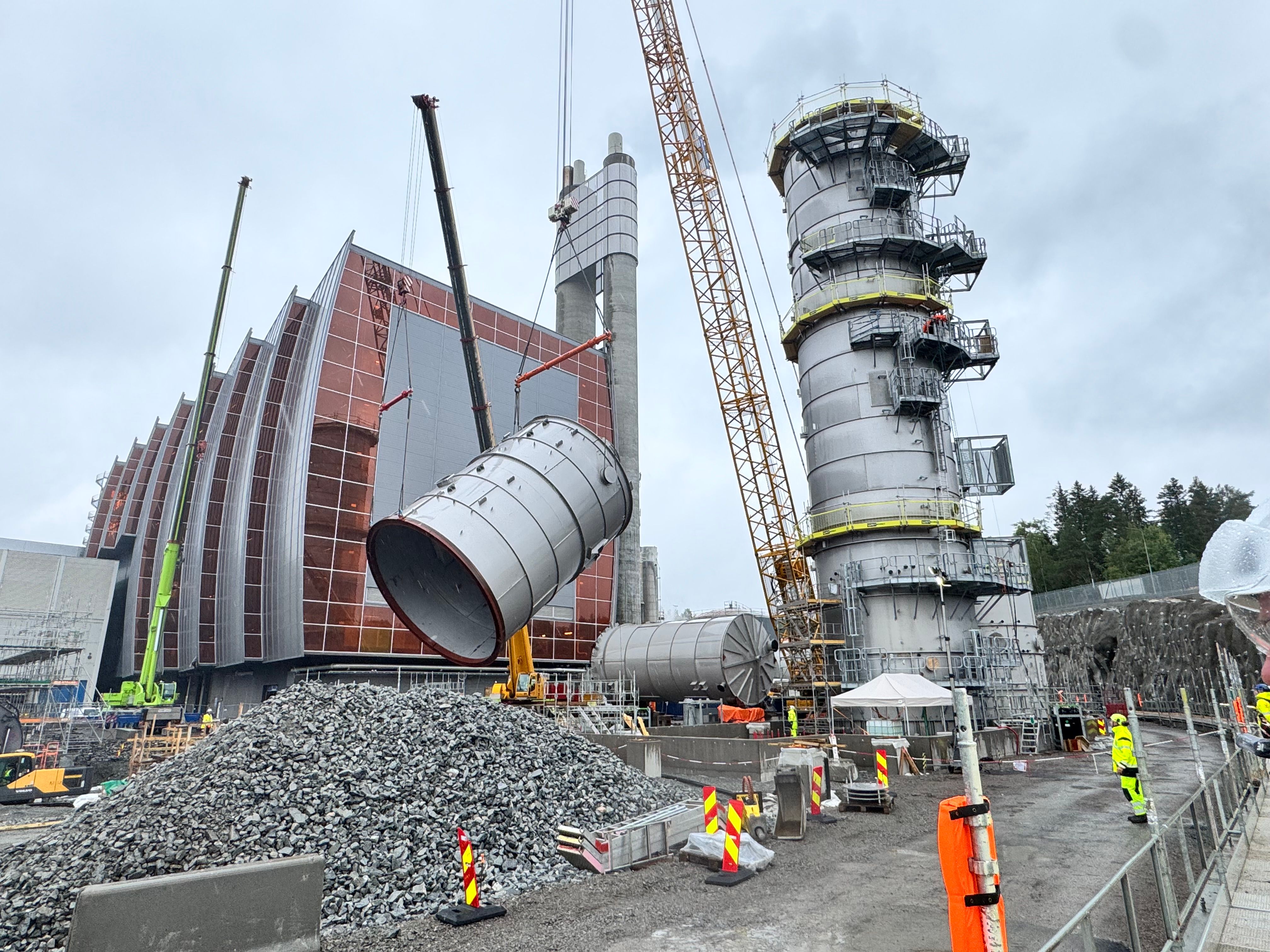 Cranes lift a large cylindrical module at a construction site with a modern building featuring a ribbed, copper-colored facade and a tall grey industrial tower.