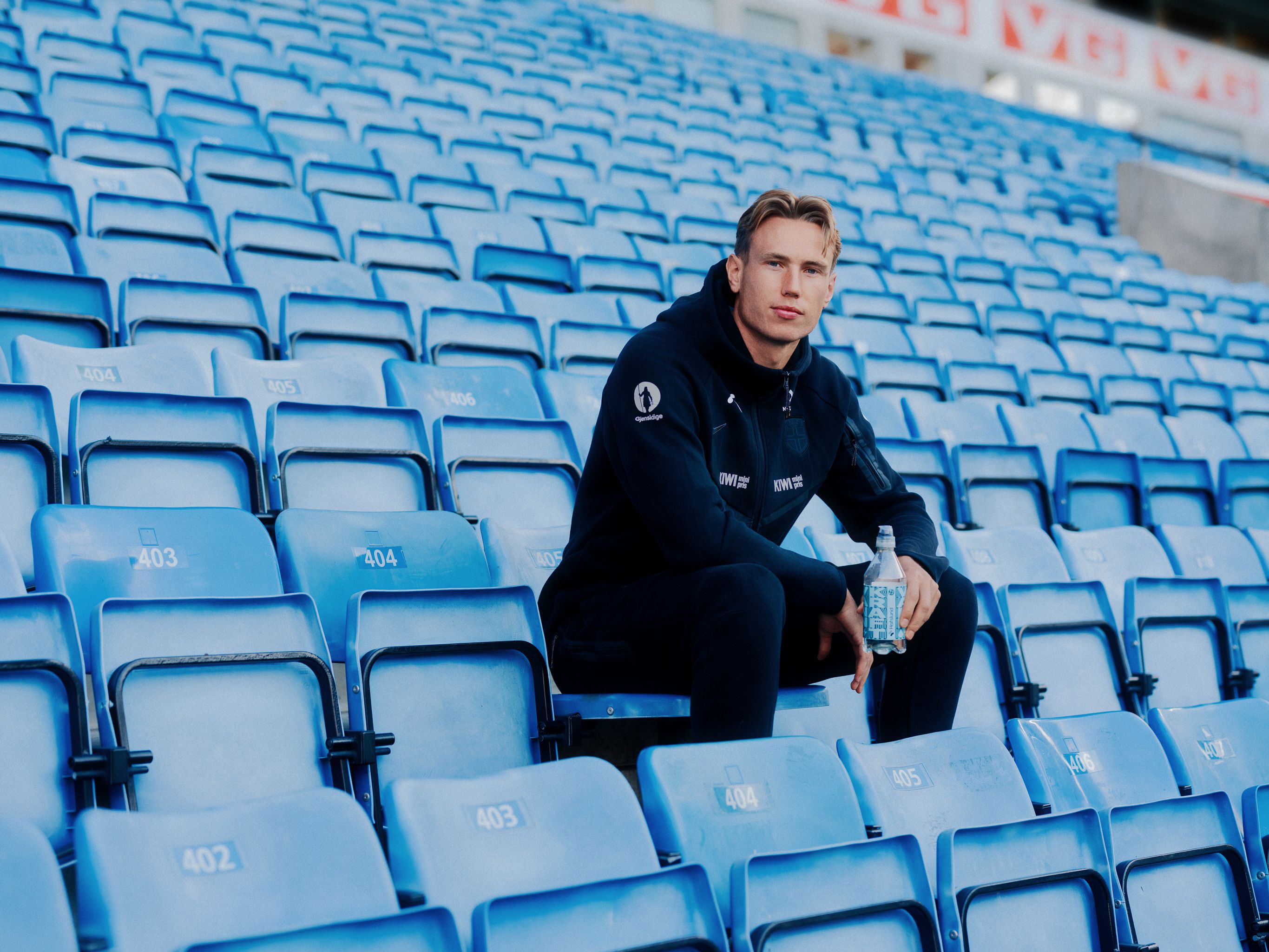 A young man in a dark tracksuit sits in an empty stadium of blue seats, holding a water bottle.