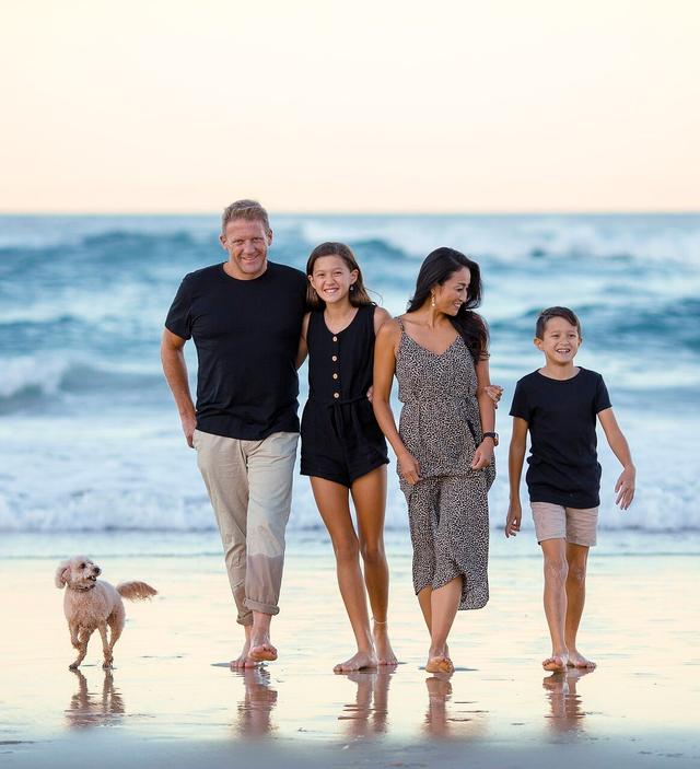 A smiling family of four and a small dog walk barefoot on a wet beach, with ocean waves in the background.