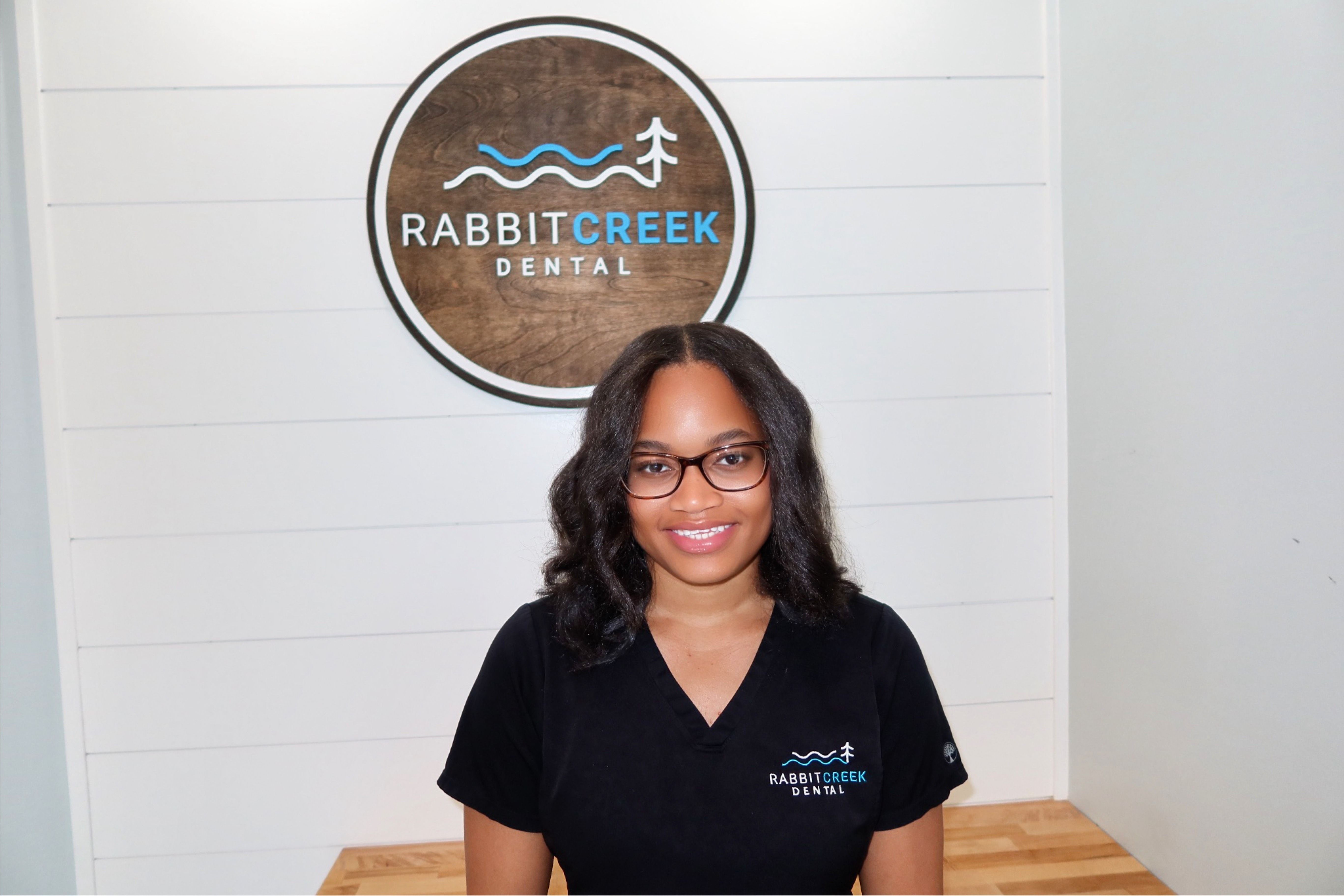 a woman wearing glasses and a black shirt is standing in front of a rabbit creek dental sign .