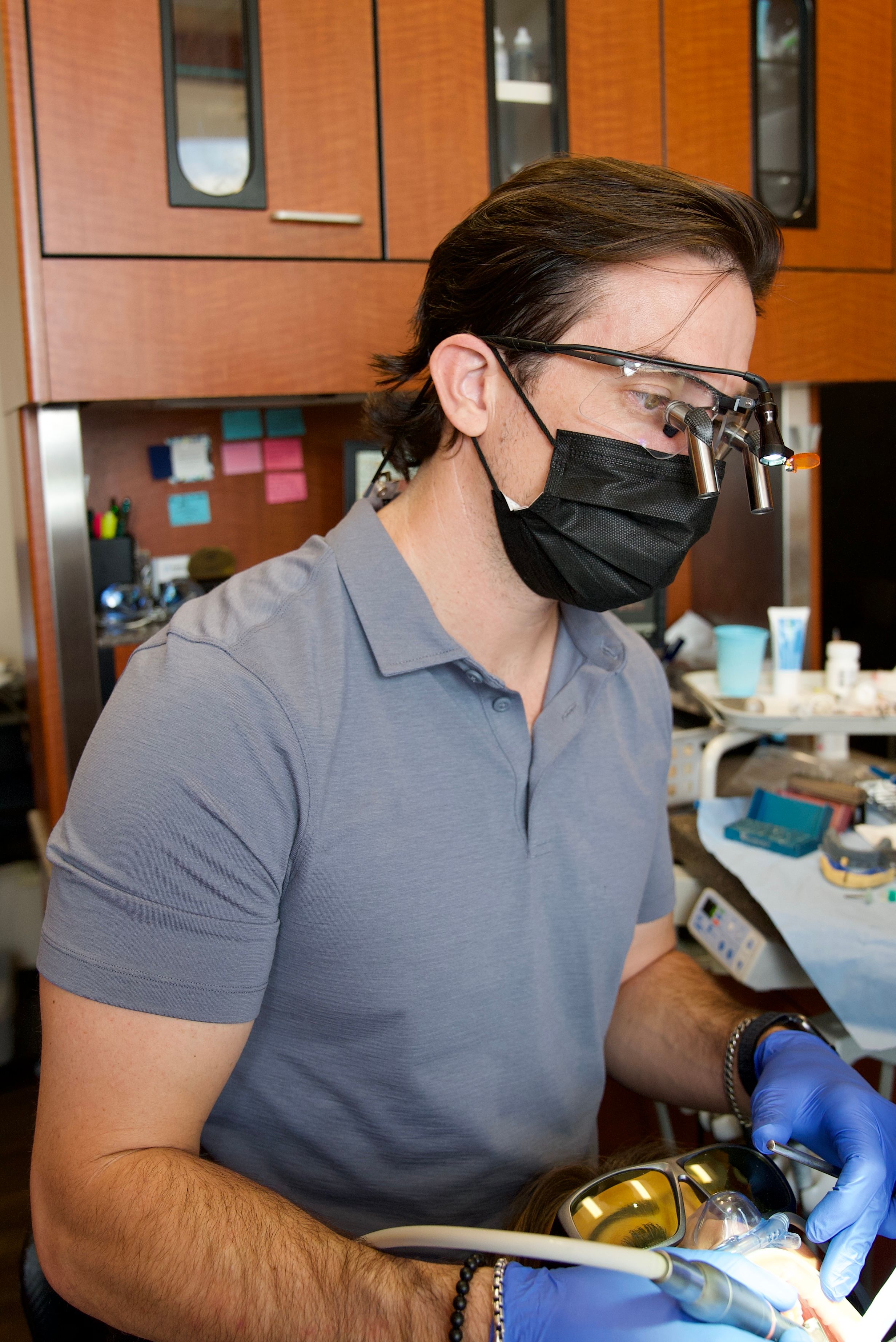 A dentist wearing a mask, magnifying loupes, and gloves performs a procedure on a patient.