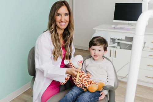 A smiling female dentist teaches a young boy to brush teeth using a giraffe plush toy with a dental model.