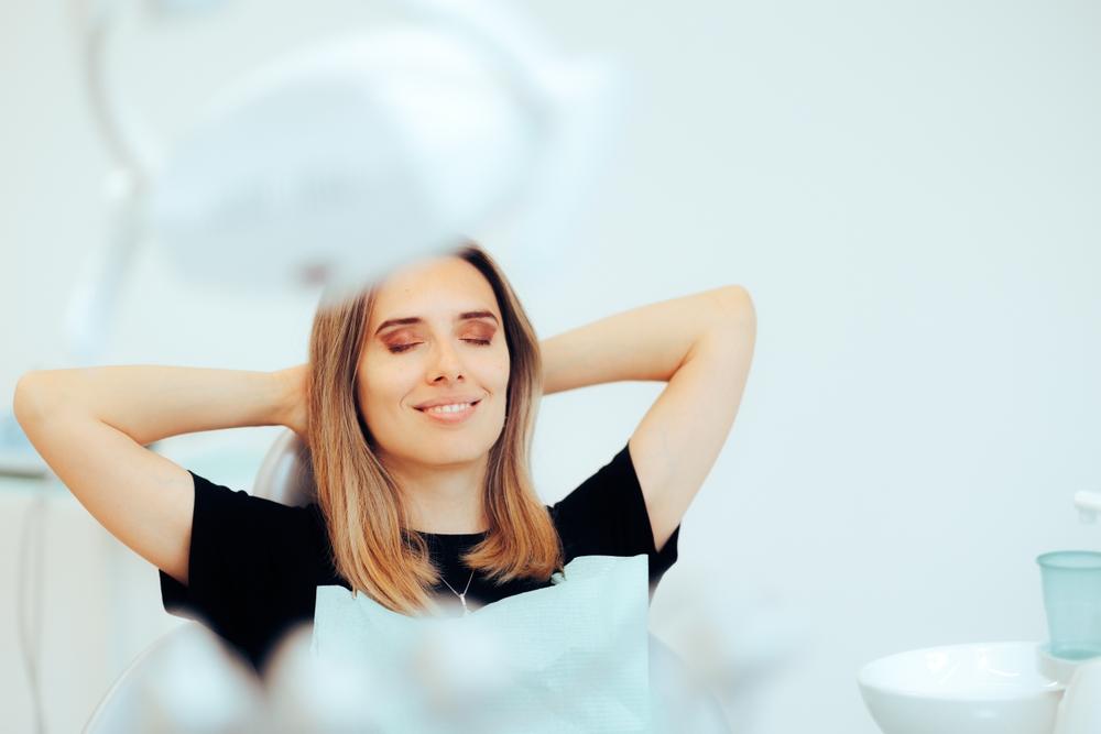 A woman relaxing in a dental chair with her eyes closed and hands behind her head.