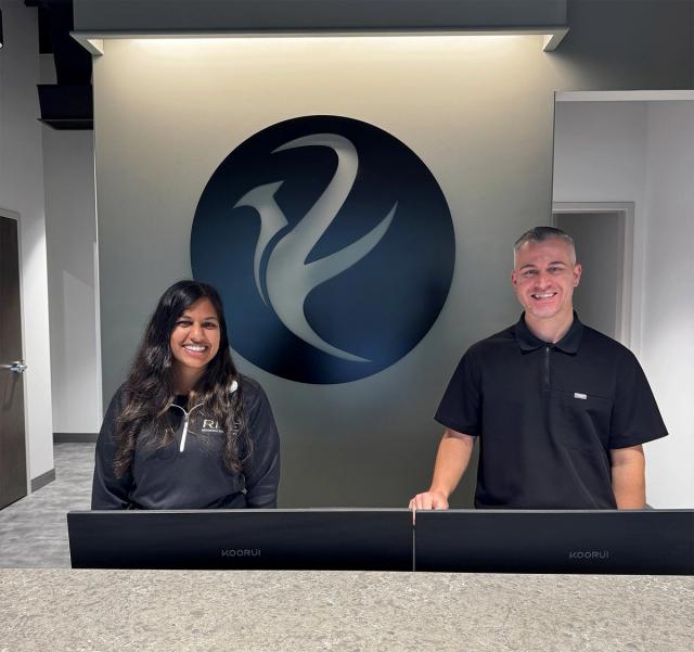 Two smiling people stand behind a reception desk with a stylized phoenix logo on the wall.