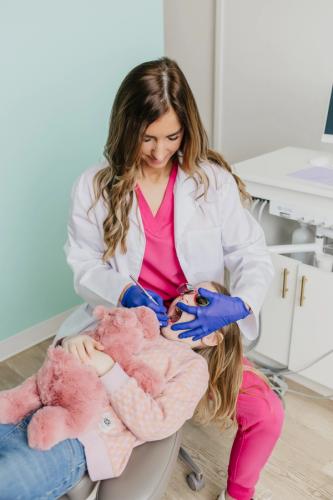 A female dentist wearing blue gloves checks a young girl's teeth. The girl lies in a dental chair holding a pink stuffed animal and wearing pink sunglasses.
