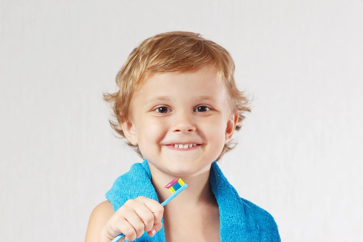Smiling child in a blue towel holding a toothbrush with toothpaste.
